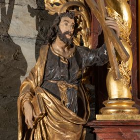 Zdjęcie nr 1: Pełnoplastyczne przedstawienie w kontrapoście ukazuje w całej postaci św. Andrzeja. Apostoł ujęty został w trzech czwartych. W uniesionej ku górze lewej ręce trzyma niewielki krzyż, określany jako krzyż św. Andrzeja. W prawej, opuszczonej wzdłuż tułowia i lekko zgiętej w łokciu, trzyma złoconą księgę zamkniętą na dwie klamry. Andrzej został ukazany jako dojrzały mężczyzna o jasnej karnacji. Ma pociągłą twarz okoloną brązowym zarostem oraz puklowane, brązowe włosy sięgające ramion. Twarz charakteryzują mocno otwarte oczy, wpatrzone w uniesiony krzyż, prosty nos oraz wydatne usta. Apostoł ubrany jest w mocno u spodu drapowaną, ciemną, luźną tunikę, wykończoną przy szyi złotą lamówką. Jest ona przewiązana w pasie złotą szarfą. Spod tuniki widać ciemne buty. Na ramionach ma narzucony złoty, mocno udrapowany płaszcz okrywający prawe ramię. Jego lewą połę przy prawym boku podtrzymuje ręką, w której trzyma księgę.   