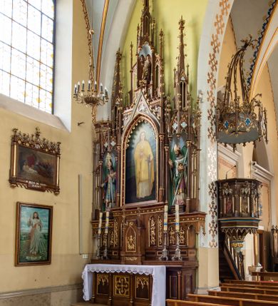 Zdjęcie nr 3: Neogotycki, przyścienny, jednokondygnacyjny ołtarz Matki Boskiej Różańcowej uzyskał architektoniczną strukturę ze zwieńczeniem. Kolorystyka ołtarza utrzymana została w tonacji ciemnego, bejcowanego drewna. Detale, ornamenty zostały wyzłocone, natomiast wypełnienia maswerków i ram wimperg są białe. Z kolei pole ramy głównego obrazu wykonano jako czerwone. Retabulum ustawiono na podwójnych gradusach zaokrąglonych od przodu. Stipes ołtarzowy uzyskał kształt prostopadłościanu z trzema czworobocznymi płycinami od frontu, ujętymi złoconymi i profilowanymi obramieniami. Płyciny rozdzielają  półkolumienki na wysokich cokolikach. Dwie skrajne płyciny stipesu wypełniono u góry maswerkiem, natomiast w środkowej znalazł się równoramienny krzyż wpisany w czteroliść. Stipes podtrzymuje drewnianą mensę z portatylem. Ołtarz przylega do wysokiego, prostopadłościennego cokołu flankowanego po bokach kolumienkami, na którym wspiera się cała nastawa. Widoczne jego partie zdobią prostokątne płyciny w obramieniach. Na cokole wspiera się predella podzielona płytkimi, prostokątnymi płycinami zakończonymi ostrołukowymi arkadkami z noskami, wypełnionymi płaskorzeźbionym i złoconym ornamentem roślinnym. Pośrodku, w przerwie między płycinami predelli, usytuowano prostopadłościenne tabernakulum w formie kapliczki, zwieńczone łukiem siodłowym, flankowane dwoma pinaklami. Ostrołukowe drzwiczki tabernakulum zdobi skrzyżowana z krzyżem kotwica oraz wyobrażenie Boskiego Serca Pana Jezusa. Poniżej widoczna jest girlanda różana. Predella wspiera trójosiową kondygnację ze zwieńczeniem. Na osi środkowej usytuowano płytką, prostokątną wnękę zakończoną ostrołukiem z arkaturą. Flankują ją smukłe kolumienki na wysokich cokolikach wspierające wysokie pinakle. Ponad wnęką widoczna jest wimperga z maswerkiem dekorowana czołgankami i kwiatonem. We wnęce umieszczono płaskorzeźbione i polichromowane przedstawienie Matki Boskiej Różańcowej ze św. Dominikiem i św. Katarzyną ze Sieny, a na zasuwie obraz ze św. Barbarą. W osiach skrajnych, pod gotyckimi baldachimami z wimpergami, rozdzielonymi pinaklami, ustawiono w niszach na postumentach figury św. Wojciecha i św. Stanisława. Święci zostali podpisani na postumentach. Konchy nisz są jasnoniebieskie natomiast ich ścianki zielone. Boki baldachimów udekorowano małymi wimpergami rozdzielonymi smukłymi pinaklami z kwiatonami i czołgankami. Baldachimy przechodzą w smukłe, ażurowe fiale dekorowane pinaklami i czołgankami oraz kwiatonami. Zwieńczenie nastawy na osi środkowej uzyskało kształt otwartej kapliczki z niszą zakończoną smukłą fialą z iglicą, dekorowaną czołgankami i kwiatonem. Po bokach kapliczki znajdują się ażurowe przypory wypełnione maswerkiem, flankowane pinaklami. W niszy umieszczono figurę św. Józefa z Dzieciątkiem. Całość retabulum flankują uszaki w formie ażurowych przypór, wypełnionych maswerkami i zdobionych parą pinakli.


