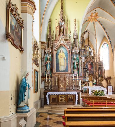 Zdjęcie nr 2: Neogotycki, przyścienny, jednokondygnacyjny ołtarz Matki Boskiej Różańcowej uzyskał architektoniczną strukturę ze zwieńczeniem. Kolorystyka ołtarza utrzymana została w tonacji ciemnego, bejcowanego drewna. Detale, ornamenty zostały wyzłocone, natomiast wypełnienia maswerków i ram wimperg są białe. Z kolei pole ramy głównego obrazu wykonano jako czerwone. Retabulum ustawiono na podwójnych gradusach zaokrąglonych od przodu. Stipes ołtarzowy uzyskał kształt prostopadłościanu z trzema czworobocznymi płycinami od frontu, ujętymi złoconymi i profilowanymi obramieniami. Płyciny rozdzielają  półkolumienki na wysokich cokolikach. Dwie skrajne płyciny stipesu wypełniono u góry maswerkiem, natomiast w środkowej znalazł się równoramienny krzyż wpisany w czteroliść. Stipes podtrzymuje drewnianą mensę z portatylem. Ołtarz przylega do wysokiego, prostopadłościennego cokołu flankowanego po bokach kolumienkami, na którym wspiera się cała nastawa. Widoczne jego partie zdobią prostokątne płyciny w obramieniach. Na cokole wspiera się predella podzielona płytkimi, prostokątnymi płycinami zakończonymi ostrołukowymi arkadkami z noskami, wypełnionymi płaskorzeźbionym i złoconym ornamentem roślinnym. Pośrodku, w przerwie między płycinami predelli, usytuowano prostopadłościenne tabernakulum w formie kapliczki, zwieńczone łukiem siodłowym, flankowane dwoma pinaklami. Ostrołukowe drzwiczki tabernakulum zdobi skrzyżowana z krzyżem kotwica oraz wyobrażenie Boskiego Serca Pana Jezusa. Poniżej widoczna jest girlanda różana. Predella wspiera trójosiową kondygnację ze zwieńczeniem. Na osi środkowej usytuowano płytką, prostokątną wnękę zakończoną ostrołukiem z arkaturą. Flankują ją smukłe kolumienki na wysokich cokolikach wspierające wysokie pinakle. Ponad wnęką widoczna jest wimperga z maswerkiem dekorowana czołgankami i kwiatonem. We wnęce umieszczono płaskorzeźbione i polichromowane przedstawienie Matki Boskiej Różańcowej ze św. Dominikiem i św. Katarzyną ze Sieny, a na zasuwie obraz ze św. Barbarą. W osiach skrajnych, pod gotyckimi baldachimami z wimpergami, rozdzielonymi pinaklami, ustawiono w niszach na postumentach figury św. Wojciecha i św. Stanisława. Święci zostali podpisani na postumentach. Konchy nisz są jasnoniebieskie natomiast ich ścianki zielone. Boki baldachimów udekorowano małymi wimpergami rozdzielonymi smukłymi pinaklami z kwiatonami i czołgankami. Baldachimy przechodzą w smukłe, ażurowe fiale dekorowane pinaklami i czołgankami oraz kwiatonami. Zwieńczenie nastawy na osi środkowej uzyskało kształt otwartej kapliczki z niszą zakończoną smukłą fialą z iglicą, dekorowaną czołgankami i kwiatonem. Po bokach kapliczki znajdują się ażurowe przypory wypełnione maswerkiem, flankowane pinaklami. W niszy umieszczono figurę św. Józefa z Dzieciątkiem. Całość retabulum flankują uszaki w formie ażurowych przypór, wypełnionych maswerkami i zdobionych parą pinakli.

