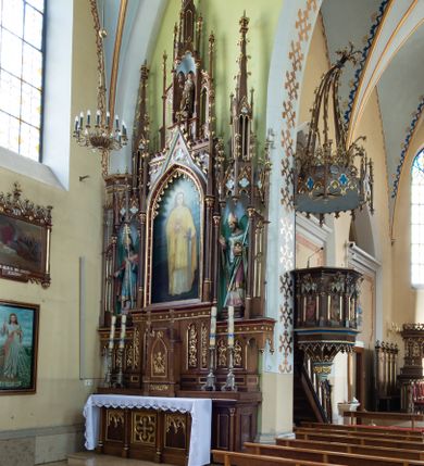 Zdjęcie nr 4: Neogotycki, przyścienny, jednokondygnacyjny ołtarz Matki Boskiej Różańcowej uzyskał architektoniczną strukturę ze zwieńczeniem. Kolorystyka ołtarza utrzymana została w tonacji ciemnego, bejcowanego drewna. Detale, ornamenty zostały wyzłocone, natomiast wypełnienia maswerków i ram wimperg są białe. Z kolei pole ramy głównego obrazu wykonano jako czerwone. Retabulum ustawiono na podwójnych gradusach zaokrąglonych od przodu. Stipes ołtarzowy uzyskał kształt prostopadłościanu z trzema czworobocznymi płycinami od frontu, ujętymi złoconymi i profilowanymi obramieniami. Płyciny rozdzielają  półkolumienki na wysokich cokolikach. Dwie skrajne płyciny stipesu wypełniono u góry maswerkiem, natomiast w środkowej znalazł się równoramienny krzyż wpisany w czteroliść. Stipes podtrzymuje drewnianą mensę z portatylem. Ołtarz przylega do wysokiego, prostopadłościennego cokołu flankowanego po bokach kolumienkami, na którym wspiera się cała nastawa. Widoczne jego partie zdobią prostokątne płyciny w obramieniach. Na cokole wspiera się predella podzielona płytkimi, prostokątnymi płycinami zakończonymi ostrołukowymi arkadkami z noskami, wypełnionymi płaskorzeźbionym i złoconym ornamentem roślinnym. Pośrodku, w przerwie między płycinami predelli, usytuowano prostopadłościenne tabernakulum w formie kapliczki, zwieńczone łukiem siodłowym, flankowane dwoma pinaklami. Ostrołukowe drzwiczki tabernakulum zdobi skrzyżowana z krzyżem kotwica oraz wyobrażenie Boskiego Serca Pana Jezusa. Poniżej widoczna jest girlanda różana. Predella wspiera trójosiową kondygnację ze zwieńczeniem. Na osi środkowej usytuowano płytką, prostokątną wnękę zakończoną ostrołukiem z arkaturą. Flankują ją smukłe kolumienki na wysokich cokolikach wspierające wysokie pinakle. Ponad wnęką widoczna jest wimperga z maswerkiem dekorowana czołgankami i kwiatonem. We wnęce umieszczono płaskorzeźbione i polichromowane przedstawienie Matki Boskiej Różańcowej ze św. Dominikiem i św. Katarzyną ze Sieny, a na zasuwie obraz ze św. Barbarą. W osiach skrajnych, pod gotyckimi baldachimami z wimpergami, rozdzielonymi pinaklami, ustawiono w niszach na postumentach figury św. Wojciecha i św. Stanisława. Święci zostali podpisani na postumentach. Konchy nisz są jasnoniebieskie natomiast ich ścianki zielone. Boki baldachimów udekorowano małymi wimpergami rozdzielonymi smukłymi pinaklami z kwiatonami i czołgankami. Baldachimy przechodzą w smukłe, ażurowe fiale dekorowane pinaklami i czołgankami oraz kwiatonami. Zwieńczenie nastawy na osi środkowej uzyskało kształt otwartej kapliczki z niszą zakończoną smukłą fialą z iglicą, dekorowaną czołgankami i kwiatonem. Po bokach kapliczki znajdują się ażurowe przypory wypełnione maswerkiem, flankowane pinaklami. W niszy umieszczono figurę św. Józefa z Dzieciątkiem. Całość retabulum flankują uszaki w formie ażurowych przypór, wypełnionych maswerkami i zdobionych parą pinakli.

