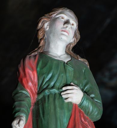 Zdjęcie nr 1: Figura ukazuje w całej postaci stojącego na płaskim postumencie w nieznacznym kontrapoście św. Jana Ewangelistę. Apostoł został przedstawiony frontalnie z nieznacznie odchyloną głową w lewą stronę. Lewą rękę zgiętą w łokciu przyciska do pasa. Drugą ma lekko odsuniętą i opuszczoną wzdłuż tułowia. Jan ma pociągłą twarz o jasnej karnacji, duży nos, wydatne usta i melancholijnie skierowane ku górze oczy. Jego głowę otaczają długie, brązowe, falujące włosy spływające na ramiona i odsłaniające uszy. Ubrany jest w zieloną tunikę przewiązaną w pasie z charakterystycznym zagięciem na piersi oraz czerwony płaszcz przerzucony przez prawe ramię, którego lewą połę przyciska ręką. Spod tuniki widoczne są fragmenty bosych stóp.