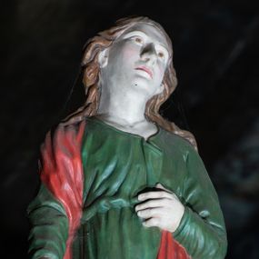 Zdjęcie nr 1: Figura ukazuje w całej postaci stojącego na płaskim postumencie w nieznacznym kontrapoście św. Jana Ewangelistę. Apostoł został przedstawiony frontalnie z nieznacznie odchyloną głową w lewą stronę. Lewą rękę zgiętą w łokciu przyciska do pasa. Drugą ma lekko odsuniętą i opuszczoną wzdłuż tułowia. Jan ma pociągłą twarz o jasnej karnacji, duży nos, wydatne usta i melancholijnie skierowane ku górze oczy. Jego głowę otaczają długie, brązowe, falujące włosy spływające na ramiona i odsłaniające uszy. Ubrany jest w zieloną tunikę przewiązaną w pasie z charakterystycznym zagięciem na piersi oraz czerwony płaszcz przerzucony przez prawe ramię, którego lewą połę przyciska ręką. Spod tuniki widoczne są fragmenty bosych stóp.