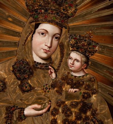 Zdjęcie nr 2: Obraz w kształcie prostokąta zbliżonego do kwadratu, ukazuje Matkę Boską z Dzieciątkiem w typie Hodegetrii krakowskiej. Drewniana, rzeźbiona i złocona sukienka wotywna przesłania obraz, odsłaniając jedynie twarze, dłonie, stopy oraz fragmenty tła. Matka Boska została ujęta frontalnie w półpostaci, trzymając na lewym przedramieniu małego Jezusa. Prawą dłonią zgiętej w łokciu ręki wskazuje na Dzieciątko, trzymając w niej równocześnie gruszkę, zaś lewą, opuszczoną, podtrzymuje jego stopy. Maria nieznacznie pochyla głowę w stronę Syna. Jej twarz ma jasną karnację o rumianych policzkach, duże, szeroko rozstawione oczy, prosty nos, wąskie usta i tylko częściowo widoczne ciemne włosy. Mały Jezus siedzi wyprostowany na lewym ramieniu Marii. Ma nieznacznie uniesioną głowę ze spojrzeniem skierowanym na matkę. Prawą ręką błogosławi, lewą zaś opiera na trzymanej na kolanach księdze Ewangelii. Jego owalna twarz o jasnej karnacji z rumianymi policzkami widoczna jest w trzech czwartych. Na głowie spod korony wotywnej widać jasne, krótkie włosy. Obie postacie okrywa drewniana, płaskorzeźbiona i złocona sukienka wotywna z koronami. Sukienka zapewne powtarza dukt maforionu Matki Boskiej oraz kształt i drapowanie długiej tuniki, która okrywa małego Jezusa. Szaty Marii dekorowane są płaskorzeźbionym ornamentem z motywem szerokolistnych gałązek z kwiatami w kształcie rozet, naśladujących kameryzacje. Malowana kameryzacja widoczna jest także na taśmach lamujących maforion oraz spodnią suknię pod szyją i na jej rękawach. Pod szyją Marii widać drewniany krzyżyk. Tunika Dzieciątka jest pokryta płaskorzeźbionymi rozetami z naśladowaniem malowanej kameryzacji. Obie postacie mają na głowach zamknięte, płaskorzeźbione i złocone korony z globem i krzyżykiem. Także one są dekorowane malowaną kameryzacją. Marię i Dzieciątko otaczają nakładane, płaskorzeźbione i złocone promienie, zakończone w jaskółczy ogon, ułożone na kształt glorii.