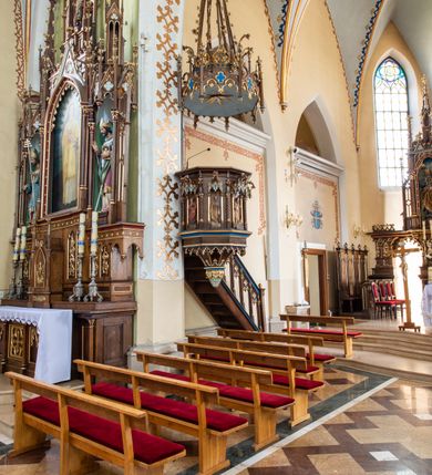 Zdjęcie nr 4: Przyścienną, wiszącą ambonę usytuowano przy łuku tęczowym na zachodniej ścianie prezbiterium. Ambona składa się z baldachimu, zredukowanego zaplecka, kosza oraz prowadzących do niej przyściennych schodów. Wykonana jest z drewna dębowego, bejcowanego i lakierowanego. Partie ornamentalne zostały wyzłocone lub też polichromowane na czerwono i niebiesko. Kosz uzyskał kształt ośmioboczny. Wieńczy go parapet w formie profilowanego gzymsu koronującego. Kolejne pięć boków kosza flankują spiralne kolumienki o liściastych kapitelach ustawionych na wysokich cokolikach, podtrzymujących imposty z pinaklami. Pole każdego boku ozdobiono maswerkowym baldachimem zwieńczonym wygiętą do przodu sterczyną zakończoną kwiatonem. Baldachimy wspierają smukłe kolumienki o liściastych kapitelach ustawione na wysokich cokolikach. Cokoliki kolumienek narożnych i tych podtrzymujących baldachimy wspierają się na łamanym cokole obiegającym cały kosz. Cokół dekorowany od spodu arkaturą złoconą i polichromowaną na czerwono i niebiesko oddziela górną część ambony od jej spodu. Spód ambony utworzono z spływów koncentrujących się na środku na ośmiobocznej, profilowanej opasce dekorowanej od góry stylizowanym motywem lilii heraldycznej, a od dołu stylizowanymi, złoconymi liśćmi winnej latorośli otaczającej dużą kiść winnego grona. Pod baldachimami na czterech bokach umieszczono wykonane w głębokim reliefie przedstawienia stojących ewangelistów. Piąty bok ambony, przylegający do schodów, zdobi zredukowany baldachim pod którym umieszczono złocony i srebrzony kwiat słonecznika. Każdy z ewangelistów stoi na niewielkim postumencie, ustawionym na głównym cokole. Na każdym postumencie widnieję imię przedstawionego ewangelisty. Na południowym boku kosza umieszczono św. Jan. Ukazany został frontalnie z głową ujętą w trzech czwartych w lewo. Ma on długie, falowane, brązowe włosy spływające na ramiona, głowę pochyloną w prawo, wzrok skierowany ku górze. W lewej ręce trzyma księgę, a w prawej, przyciśniętej do piersi pióro. Ubrany jest w długą zieloną tunikę, nakrytą czerwonym płaszczem. Po lewej stronie, u jego stóp widnieje czarny orzeł. Na kolejnym boku znajduje się św. Marek. Wyobrażono go frontalnie z prawą ręką w geście błogosławieństwa, natomiast w lewej trzyma rotulus. Ma on krótkie, brązowe włosy oraz brodę. Ubrany jest w niebieską tunikę odsłaniającą fragmenty bosych stóp i żółty płaszcz. Po lewej stronie przy jego nogach znajduje się lew. Na następnym boku ukazano frontalnie stojącego św. Mateusza. Ewangelista w lewej ręce przyciśniętej do piersi trzyma rotulus, a prawą opuszczoną wzdłóż tułowia wyciąga do przodu. Ma długie, brązowe, falowane włosy spływające na kark oraz brodę. Ubrany jest w blado różową tunikę i żółty płaszcz. Przy jego lewej stopie znajduje się uskrzydlona anielska główka. Dalej znajduje się także przedstawiony frontalnie św. Łukasz. Równocześnie prawą i lewą ręką podtrzymuje zamknietą księgę. Ma on łysiejące czoło i głowę otoczoną siwiejącymi włosami i brodą. Ewangelista ma na sobie brązową tunikę u spodu odsłaniającą fragmenty bosych stóp oraz bladoniebieski płaszcz przerzucony przez prawe ramię. U jego stóp, po lewej stronie, znajduje się wół. Na ambonę prowadzą ulokowane przy ścianie schody o dziesięciu stopniach. Ich balustradę artykułują kolumienki o liściastych kapitelach flankujące ażurowe, ostrołukowe arkady wypełnione u góry złoconym maswerkiem. Zaplecek ambony został zredukowany do szerokiej listwy dekorowanej stylizowaną ostrołukową arkadką. W jej górnej części wyobrażono dwie tablice Dekalogu z numerami przykazań: na jednej: I, II, III, IV, V; na drugiej: VI, VII, VIII, IX, X. Ponad amboną zawieszono wieloboczny baldachim zwieńczony ażurową wiązką spiętych razem pierścieniem zdobionych czołgankami prętów, tworzących dekoracyjny kwiaton wygięty do przodu. Boki baldachimu ozdobiono wycinanymi w desce maswerkami flankowanymi pinaklami, zwieńczonymi powyginanymi sterczynami. Niebieskie podniebie baldachimu zdobi srebrzone wyobrażenie gołębicy Ducha Świętego na tle złocistych promieni. 