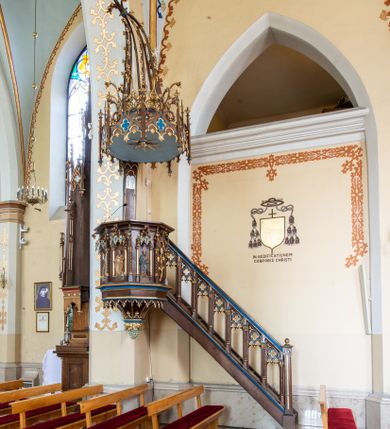 Zdjęcie nr 1: Przyścienną, wiszącą ambonę usytuowano przy łuku tęczowym na zachodniej ścianie prezbiterium. Ambona składa się z baldachimu, zredukowanego zaplecka, kosza oraz prowadzących do niej przyściennych schodów. Wykonana jest z drewna dębowego, bejcowanego i lakierowanego. Partie ornamentalne zostały wyzłocone lub też polichromowane na czerwono i niebiesko. Kosz uzyskał kształt ośmioboczny. Wieńczy go parapet w formie profilowanego gzymsu koronującego. Kolejne pięć boków kosza flankują spiralne kolumienki o liściastych kapitelach ustawionych na wysokich cokolikach, podtrzymujących imposty z pinaklami. Pole każdego boku ozdobiono maswerkowym baldachimem zwieńczonym wygiętą do przodu sterczyną zakończoną kwiatonem. Baldachimy wspierają smukłe kolumienki o liściastych kapitelach ustawione na wysokich cokolikach. Cokoliki kolumienek narożnych i tych podtrzymujących baldachimy wspierają się na łamanym cokole obiegającym cały kosz. Cokół dekorowany od spodu arkaturą złoconą i polichromowaną na czerwono i niebiesko oddziela górną część ambony od jej spodu. Spód ambony utworzono z spływów koncentrujących się na środku na ośmiobocznej, profilowanej opasce dekorowanej od góry stylizowanym motywem lilii heraldycznej, a od dołu stylizowanymi, złoconymi liśćmi winnej latorośli otaczającej dużą kiść winnego grona. Pod baldachimami na czterech bokach umieszczono wykonane w głębokim reliefie przedstawienia stojących ewangelistów. Piąty bok ambony, przylegający do schodów, zdobi zredukowany baldachim pod którym umieszczono złocony i srebrzony kwiat słonecznika. Każdy z ewangelistów stoi na niewielkim postumencie, ustawionym na głównym cokole. Na każdym postumencie widnieję imię przedstawionego ewangelisty. Na południowym boku kosza umieszczono św. Jan. Ukazany został frontalnie z głową ujętą w trzech czwartych w lewo. Ma on długie, falowane, brązowe włosy spływające na ramiona, głowę pochyloną w prawo, wzrok skierowany ku górze. W lewej ręce trzyma księgę, a w prawej, przyciśniętej do piersi pióro. Ubrany jest w długą zieloną tunikę, nakrytą czerwonym płaszczem. Po lewej stronie, u jego stóp widnieje czarny orzeł. Na kolejnym boku znajduje się św. Marek. Wyobrażono go frontalnie z prawą ręką w geście błogosławieństwa, natomiast w lewej trzyma rotulus. Ma on krótkie, brązowe włosy oraz brodę. Ubrany jest w niebieską tunikę odsłaniającą fragmenty bosych stóp i żółty płaszcz. Po lewej stronie przy jego nogach znajduje się lew. Na następnym boku ukazano frontalnie stojącego św. Mateusza. Ewangelista w lewej ręce przyciśniętej do piersi trzyma rotulus, a prawą opuszczoną wzdłóż tułowia wyciąga do przodu. Ma długie, brązowe, falowane włosy spływające na kark oraz brodę. Ubrany jest w blado różową tunikę i żółty płaszcz. Przy jego lewej stopie znajduje się uskrzydlona anielska główka. Dalej znajduje się także przedstawiony frontalnie św. Łukasz. Równocześnie prawą i lewą ręką podtrzymuje zamknietą księgę. Ma on łysiejące czoło i głowę otoczoną siwiejącymi włosami i brodą. Ewangelista ma na sobie brązową tunikę u spodu odsłaniającą fragmenty bosych stóp oraz bladoniebieski płaszcz przerzucony przez prawe ramię. U jego stóp, po lewej stronie, znajduje się wół. Na ambonę prowadzą ulokowane przy ścianie schody o dziesięciu stopniach. Ich balustradę artykułują kolumienki o liściastych kapitelach flankujące ażurowe, ostrołukowe arkady wypełnione u góry złoconym maswerkiem. Zaplecek ambony został zredukowany do szerokiej listwy dekorowanej stylizowaną ostrołukową arkadką. W jej górnej części wyobrażono dwie tablice Dekalogu z numerami przykazań: na jednej: I, II, III, IV, V; na drugiej: VI, VII, VIII, IX, X. Ponad amboną zawieszono wieloboczny baldachim zwieńczony ażurową wiązką spiętych razem pierścieniem zdobionych czołgankami prętów, tworzących dekoracyjny kwiaton wygięty do przodu. Boki baldachimu ozdobiono wycinanymi w desce maswerkami flankowanymi pinaklami, zwieńczonymi powyginanymi sterczynami. Niebieskie podniebie baldachimu zdobi srebrzone wyobrażenie gołębicy Ducha Świętego na tle złocistych promieni. 