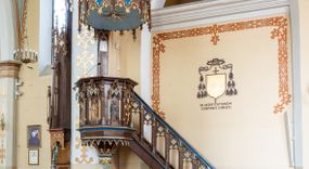 Zdjęcie nr 1: Przyścienną, wiszącą ambonę usytuowano przy łuku tęczowym na zachodniej ścianie prezbiterium. Ambona składa się z baldachimu, zredukowanego zaplecka, kosza oraz prowadzących do niej przyściennych schodów. Wykonana jest z drewna dębowego, bejcowanego i lakierowanego. Partie ornamentalne zostały wyzłocone lub też polichromowane na czerwono i niebiesko. Kosz uzyskał kształt ośmioboczny. Wieńczy go parapet w formie profilowanego gzymsu koronującego. Kolejne pięć boków kosza flankują spiralne kolumienki o liściastych kapitelach ustawionych na wysokich cokolikach, podtrzymujących imposty z pinaklami. Pole każdego boku ozdobiono maswerkowym baldachimem zwieńczonym wygiętą do przodu sterczyną zakończoną kwiatonem. Baldachimy wspierają smukłe kolumienki o liściastych kapitelach ustawione na wysokich cokolikach. Cokoliki kolumienek narożnych i tych podtrzymujących baldachimy wspierają się na łamanym cokole obiegającym cały kosz. Cokół dekorowany od spodu arkaturą złoconą i polichromowaną na czerwono i niebiesko oddziela górną część ambony od jej spodu. Spód ambony utworzono z spływów koncentrujących się na środku na ośmiobocznej, profilowanej opasce dekorowanej od góry stylizowanym motywem lilii heraldycznej, a od dołu stylizowanymi, złoconymi liśćmi winnej latorośli otaczającej dużą kiść winnego grona. Pod baldachimami na czterech bokach umieszczono wykonane w głębokim reliefie przedstawienia stojących ewangelistów. Piąty bok ambony, przylegający do schodów, zdobi zredukowany baldachim pod którym umieszczono złocony i srebrzony kwiat słonecznika. Każdy z ewangelistów stoi na niewielkim postumencie, ustawionym na głównym cokole. Na każdym postumencie widnieję imię przedstawionego ewangelisty. Na południowym boku kosza umieszczono św. Jan. Ukazany został frontalnie z głową ujętą w trzech czwartych w lewo. Ma on długie, falowane, brązowe włosy spływające na ramiona, głowę pochyloną w prawo, wzrok skierowany ku górze. W lewej ręce trzyma księgę, a w prawej, przyciśniętej do piersi pióro. Ubrany jest w długą zieloną tunikę, nakrytą czerwonym płaszczem. Po lewej stronie, u jego stóp widnieje czarny orzeł. Na kolejnym boku znajduje się św. Marek. Wyobrażono go frontalnie z prawą ręką w geście błogosławieństwa, natomiast w lewej trzyma rotulus. Ma on krótkie, brązowe włosy oraz brodę. Ubrany jest w niebieską tunikę odsłaniającą fragmenty bosych stóp i żółty płaszcz. Po lewej stronie przy jego nogach znajduje się lew. Na następnym boku ukazano frontalnie stojącego św. Mateusza. Ewangelista w lewej ręce przyciśniętej do piersi trzyma rotulus, a prawą opuszczoną wzdłóż tułowia wyciąga do przodu. Ma długie, brązowe, falowane włosy spływające na kark oraz brodę. Ubrany jest w blado różową tunikę i żółty płaszcz. Przy jego lewej stopie znajduje się uskrzydlona anielska główka. Dalej znajduje się także przedstawiony frontalnie św. Łukasz. Równocześnie prawą i lewą ręką podtrzymuje zamknietą księgę. Ma on łysiejące czoło i głowę otoczoną siwiejącymi włosami i brodą. Ewangelista ma na sobie brązową tunikę u spodu odsłaniającą fragmenty bosych stóp oraz bladoniebieski płaszcz przerzucony przez prawe ramię. U jego stóp, po lewej stronie, znajduje się wół. Na ambonę prowadzą ulokowane przy ścianie schody o dziesięciu stopniach. Ich balustradę artykułują kolumienki o liściastych kapitelach flankujące ażurowe, ostrołukowe arkady wypełnione u góry złoconym maswerkiem. Zaplecek ambony został zredukowany do szerokiej listwy dekorowanej stylizowaną ostrołukową arkadką. W jej górnej części wyobrażono dwie tablice Dekalogu z numerami przykazań: na jednej: I, II, III, IV, V; na drugiej: VI, VII, VIII, IX, X. Ponad amboną zawieszono wieloboczny baldachim zwieńczony ażurową wiązką spiętych razem pierścieniem zdobionych czołgankami prętów, tworzących dekoracyjny kwiaton wygięty do przodu. Boki baldachimu ozdobiono wycinanymi w desce maswerkami flankowanymi pinaklami, zwieńczonymi powyginanymi sterczynami. Niebieskie podniebie baldachimu zdobi srebrzone wyobrażenie gołębicy Ducha Świętego na tle złocistych promieni. 