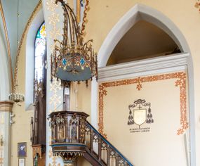 Zdjęcie nr 1: Przyścienną, wiszącą ambonę usytuowano przy łuku tęczowym na zachodniej ścianie prezbiterium. Ambona składa się z baldachimu, zredukowanego zaplecka, kosza oraz prowadzących do niej przyściennych schodów. Wykonana jest z drewna dębowego, bejcowanego i lakierowanego. Partie ornamentalne zostały wyzłocone lub też polichromowane na czerwono i niebiesko. Kosz uzyskał kształt ośmioboczny. Wieńczy go parapet w formie profilowanego gzymsu koronującego. Kolejne pięć boków kosza flankują spiralne kolumienki o liściastych kapitelach ustawionych na wysokich cokolikach, podtrzymujących imposty z pinaklami. Pole każdego boku ozdobiono maswerkowym baldachimem zwieńczonym wygiętą do przodu sterczyną zakończoną kwiatonem. Baldachimy wspierają smukłe kolumienki o liściastych kapitelach ustawione na wysokich cokolikach. Cokoliki kolumienek narożnych i tych podtrzymujących baldachimy wspierają się na łamanym cokole obiegającym cały kosz. Cokół dekorowany od spodu arkaturą złoconą i polichromowaną na czerwono i niebiesko oddziela górną część ambony od jej spodu. Spód ambony utworzono z spływów koncentrujących się na środku na ośmiobocznej, profilowanej opasce dekorowanej od góry stylizowanym motywem lilii heraldycznej, a od dołu stylizowanymi, złoconymi liśćmi winnej latorośli otaczającej dużą kiść winnego grona. Pod baldachimami na czterech bokach umieszczono wykonane w głębokim reliefie przedstawienia stojących ewangelistów. Piąty bok ambony, przylegający do schodów, zdobi zredukowany baldachim pod którym umieszczono złocony i srebrzony kwiat słonecznika. Każdy z ewangelistów stoi na niewielkim postumencie, ustawionym na głównym cokole. Na każdym postumencie widnieję imię przedstawionego ewangelisty. Na południowym boku kosza umieszczono św. Jan. Ukazany został frontalnie z głową ujętą w trzech czwartych w lewo. Ma on długie, falowane, brązowe włosy spływające na ramiona, głowę pochyloną w prawo, wzrok skierowany ku górze. W lewej ręce trzyma księgę, a w prawej, przyciśniętej do piersi pióro. Ubrany jest w długą zieloną tunikę, nakrytą czerwonym płaszczem. Po lewej stronie, u jego stóp widnieje czarny orzeł. Na kolejnym boku znajduje się św. Marek. Wyobrażono go frontalnie z prawą ręką w geście błogosławieństwa, natomiast w lewej trzyma rotulus. Ma on krótkie, brązowe włosy oraz brodę. Ubrany jest w niebieską tunikę odsłaniającą fragmenty bosych stóp i żółty płaszcz. Po lewej stronie przy jego nogach znajduje się lew. Na następnym boku ukazano frontalnie stojącego św. Mateusza. Ewangelista w lewej ręce przyciśniętej do piersi trzyma rotulus, a prawą opuszczoną wzdłóż tułowia wyciąga do przodu. Ma długie, brązowe, falowane włosy spływające na kark oraz brodę. Ubrany jest w blado różową tunikę i żółty płaszcz. Przy jego lewej stopie znajduje się uskrzydlona anielska główka. Dalej znajduje się także przedstawiony frontalnie św. Łukasz. Równocześnie prawą i lewą ręką podtrzymuje zamknietą księgę. Ma on łysiejące czoło i głowę otoczoną siwiejącymi włosami i brodą. Ewangelista ma na sobie brązową tunikę u spodu odsłaniającą fragmenty bosych stóp oraz bladoniebieski płaszcz przerzucony przez prawe ramię. U jego stóp, po lewej stronie, znajduje się wół. Na ambonę prowadzą ulokowane przy ścianie schody o dziesięciu stopniach. Ich balustradę artykułują kolumienki o liściastych kapitelach flankujące ażurowe, ostrołukowe arkady wypełnione u góry złoconym maswerkiem. Zaplecek ambony został zredukowany do szerokiej listwy dekorowanej stylizowaną ostrołukową arkadką. W jej górnej części wyobrażono dwie tablice Dekalogu z numerami przykazań: na jednej: I, II, III, IV, V; na drugiej: VI, VII, VIII, IX, X. Ponad amboną zawieszono wieloboczny baldachim zwieńczony ażurową wiązką spiętych razem pierścieniem zdobionych czołgankami prętów, tworzących dekoracyjny kwiaton wygięty do przodu. Boki baldachimu ozdobiono wycinanymi w desce maswerkami flankowanymi pinaklami, zwieńczonymi powyginanymi sterczynami. Niebieskie podniebie baldachimu zdobi srebrzone wyobrażenie gołębicy Ducha Świętego na tle złocistych promieni. 