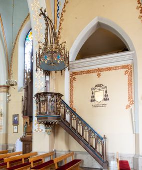 Zdjęcie nr 1: Przyścienną, wiszącą ambonę usytuowano przy łuku tęczowym na zachodniej ścianie prezbiterium. Ambona składa się z baldachimu, zredukowanego zaplecka, kosza oraz prowadzących do niej przyściennych schodów. Wykonana jest z drewna dębowego, bejcowanego i lakierowanego. Partie ornamentalne zostały wyzłocone lub też polichromowane na czerwono i niebiesko. Kosz uzyskał kształt ośmioboczny. Wieńczy go parapet w formie profilowanego gzymsu koronującego. Kolejne pięć boków kosza flankują spiralne kolumienki o liściastych kapitelach ustawionych na wysokich cokolikach, podtrzymujących imposty z pinaklami. Pole każdego boku ozdobiono maswerkowym baldachimem zwieńczonym wygiętą do przodu sterczyną zakończoną kwiatonem. Baldachimy wspierają smukłe kolumienki o liściastych kapitelach ustawione na wysokich cokolikach. Cokoliki kolumienek narożnych i tych podtrzymujących baldachimy wspierają się na łamanym cokole obiegającym cały kosz. Cokół dekorowany od spodu arkaturą złoconą i polichromowaną na czerwono i niebiesko oddziela górną część ambony od jej spodu. Spód ambony utworzono z spływów koncentrujących się na środku na ośmiobocznej, profilowanej opasce dekorowanej od góry stylizowanym motywem lilii heraldycznej, a od dołu stylizowanymi, złoconymi liśćmi winnej latorośli otaczającej dużą kiść winnego grona. Pod baldachimami na czterech bokach umieszczono wykonane w głębokim reliefie przedstawienia stojących ewangelistów. Piąty bok ambony, przylegający do schodów, zdobi zredukowany baldachim pod którym umieszczono złocony i srebrzony kwiat słonecznika. Każdy z ewangelistów stoi na niewielkim postumencie, ustawionym na głównym cokole. Na każdym postumencie widnieję imię przedstawionego ewangelisty. Na południowym boku kosza umieszczono św. Jan. Ukazany został frontalnie z głową ujętą w trzech czwartych w lewo. Ma on długie, falowane, brązowe włosy spływające na ramiona, głowę pochyloną w prawo, wzrok skierowany ku górze. W lewej ręce trzyma księgę, a w prawej, przyciśniętej do piersi pióro. Ubrany jest w długą zieloną tunikę, nakrytą czerwonym płaszczem. Po lewej stronie, u jego stóp widnieje czarny orzeł. Na kolejnym boku znajduje się św. Marek. Wyobrażono go frontalnie z prawą ręką w geście błogosławieństwa, natomiast w lewej trzyma rotulus. Ma on krótkie, brązowe włosy oraz brodę. Ubrany jest w niebieską tunikę odsłaniającą fragmenty bosych stóp i żółty płaszcz. Po lewej stronie przy jego nogach znajduje się lew. Na następnym boku ukazano frontalnie stojącego św. Mateusza. Ewangelista w lewej ręce przyciśniętej do piersi trzyma rotulus, a prawą opuszczoną wzdłóż tułowia wyciąga do przodu. Ma długie, brązowe, falowane włosy spływające na kark oraz brodę. Ubrany jest w blado różową tunikę i żółty płaszcz. Przy jego lewej stopie znajduje się uskrzydlona anielska główka. Dalej znajduje się także przedstawiony frontalnie św. Łukasz. Równocześnie prawą i lewą ręką podtrzymuje zamknietą księgę. Ma on łysiejące czoło i głowę otoczoną siwiejącymi włosami i brodą. Ewangelista ma na sobie brązową tunikę u spodu odsłaniającą fragmenty bosych stóp oraz bladoniebieski płaszcz przerzucony przez prawe ramię. U jego stóp, po lewej stronie, znajduje się wół. Na ambonę prowadzą ulokowane przy ścianie schody o dziesięciu stopniach. Ich balustradę artykułują kolumienki o liściastych kapitelach flankujące ażurowe, ostrołukowe arkady wypełnione u góry złoconym maswerkiem. Zaplecek ambony został zredukowany do szerokiej listwy dekorowanej stylizowaną ostrołukową arkadką. W jej górnej części wyobrażono dwie tablice Dekalogu z numerami przykazań: na jednej: I, II, III, IV, V; na drugiej: VI, VII, VIII, IX, X. Ponad amboną zawieszono wieloboczny baldachim zwieńczony ażurową wiązką spiętych razem pierścieniem zdobionych czołgankami prętów, tworzących dekoracyjny kwiaton wygięty do przodu. Boki baldachimu ozdobiono wycinanymi w desce maswerkami flankowanymi pinaklami, zwieńczonymi powyginanymi sterczynami. Niebieskie podniebie baldachimu zdobi srebrzone wyobrażenie gołębicy Ducha Świętego na tle złocistych promieni. 