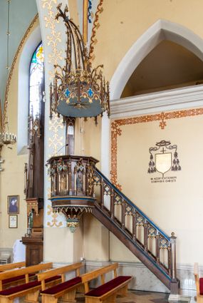 Zdjęcie nr 1: Przyścienną, wiszącą ambonę usytuowano przy łuku tęczowym na zachodniej ścianie prezbiterium. Ambona składa się z baldachimu, zredukowanego zaplecka, kosza oraz prowadzących do niej przyściennych schodów. Wykonana jest z drewna dębowego, bejcowanego i lakierowanego. Partie ornamentalne zostały wyzłocone lub też polichromowane na czerwono i niebiesko. Kosz uzyskał kształt ośmioboczny. Wieńczy go parapet w formie profilowanego gzymsu koronującego. Kolejne pięć boków kosza flankują spiralne kolumienki o liściastych kapitelach ustawionych na wysokich cokolikach, podtrzymujących imposty z pinaklami. Pole każdego boku ozdobiono maswerkowym baldachimem zwieńczonym wygiętą do przodu sterczyną zakończoną kwiatonem. Baldachimy wspierają smukłe kolumienki o liściastych kapitelach ustawione na wysokich cokolikach. Cokoliki kolumienek narożnych i tych podtrzymujących baldachimy wspierają się na łamanym cokole obiegającym cały kosz. Cokół dekorowany od spodu arkaturą złoconą i polichromowaną na czerwono i niebiesko oddziela górną część ambony od jej spodu. Spód ambony utworzono z spływów koncentrujących się na środku na ośmiobocznej, profilowanej opasce dekorowanej od góry stylizowanym motywem lilii heraldycznej, a od dołu stylizowanymi, złoconymi liśćmi winnej latorośli otaczającej dużą kiść winnego grona. Pod baldachimami na czterech bokach umieszczono wykonane w głębokim reliefie przedstawienia stojących ewangelistów. Piąty bok ambony, przylegający do schodów, zdobi zredukowany baldachim pod którym umieszczono złocony i srebrzony kwiat słonecznika. Każdy z ewangelistów stoi na niewielkim postumencie, ustawionym na głównym cokole. Na każdym postumencie widnieję imię przedstawionego ewangelisty. Na południowym boku kosza umieszczono św. Jan. Ukazany został frontalnie z głową ujętą w trzech czwartych w lewo. Ma on długie, falowane, brązowe włosy spływające na ramiona, głowę pochyloną w prawo, wzrok skierowany ku górze. W lewej ręce trzyma księgę, a w prawej, przyciśniętej do piersi pióro. Ubrany jest w długą zieloną tunikę, nakrytą czerwonym płaszczem. Po lewej stronie, u jego stóp widnieje czarny orzeł. Na kolejnym boku znajduje się św. Marek. Wyobrażono go frontalnie z prawą ręką w geście błogosławieństwa, natomiast w lewej trzyma rotulus. Ma on krótkie, brązowe włosy oraz brodę. Ubrany jest w niebieską tunikę odsłaniającą fragmenty bosych stóp i żółty płaszcz. Po lewej stronie przy jego nogach znajduje się lew. Na następnym boku ukazano frontalnie stojącego św. Mateusza. Ewangelista w lewej ręce przyciśniętej do piersi trzyma rotulus, a prawą opuszczoną wzdłóż tułowia wyciąga do przodu. Ma długie, brązowe, falowane włosy spływające na kark oraz brodę. Ubrany jest w blado różową tunikę i żółty płaszcz. Przy jego lewej stopie znajduje się uskrzydlona anielska główka. Dalej znajduje się także przedstawiony frontalnie św. Łukasz. Równocześnie prawą i lewą ręką podtrzymuje zamknietą księgę. Ma on łysiejące czoło i głowę otoczoną siwiejącymi włosami i brodą. Ewangelista ma na sobie brązową tunikę u spodu odsłaniającą fragmenty bosych stóp oraz bladoniebieski płaszcz przerzucony przez prawe ramię. U jego stóp, po lewej stronie, znajduje się wół. Na ambonę prowadzą ulokowane przy ścianie schody o dziesięciu stopniach. Ich balustradę artykułują kolumienki o liściastych kapitelach flankujące ażurowe, ostrołukowe arkady wypełnione u góry złoconym maswerkiem. Zaplecek ambony został zredukowany do szerokiej listwy dekorowanej stylizowaną ostrołukową arkadką. W jej górnej części wyobrażono dwie tablice Dekalogu z numerami przykazań: na jednej: I, II, III, IV, V; na drugiej: VI, VII, VIII, IX, X. Ponad amboną zawieszono wieloboczny baldachim zwieńczony ażurową wiązką spiętych razem pierścieniem zdobionych czołgankami prętów, tworzących dekoracyjny kwiaton wygięty do przodu. Boki baldachimu ozdobiono wycinanymi w desce maswerkami flankowanymi pinaklami, zwieńczonymi powyginanymi sterczynami. Niebieskie podniebie baldachimu zdobi srebrzone wyobrażenie gołębicy Ducha Świętego na tle złocistych promieni. 