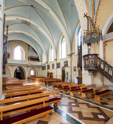 Zdjęcie nr 5: Przyścienną, wiszącą ambonę usytuowano przy łuku tęczowym na zachodniej ścianie prezbiterium. Ambona składa się z baldachimu, zredukowanego zaplecka, kosza oraz prowadzących do niej przyściennych schodów. Wykonana jest z drewna dębowego, bejcowanego i lakierowanego. Partie ornamentalne zostały wyzłocone lub też polichromowane na czerwono i niebiesko. Kosz uzyskał kształt ośmioboczny. Wieńczy go parapet w formie profilowanego gzymsu koronującego. Kolejne pięć boków kosza flankują spiralne kolumienki o liściastych kapitelach ustawionych na wysokich cokolikach, podtrzymujących imposty z pinaklami. Pole każdego boku ozdobiono maswerkowym baldachimem zwieńczonym wygiętą do przodu sterczyną zakończoną kwiatonem. Baldachimy wspierają smukłe kolumienki o liściastych kapitelach ustawione na wysokich cokolikach. Cokoliki kolumienek narożnych i tych podtrzymujących baldachimy wspierają się na łamanym cokole obiegającym cały kosz. Cokół dekorowany od spodu arkaturą złoconą i polichromowaną na czerwono i niebiesko oddziela górną część ambony od jej spodu. Spód ambony utworzono z spływów koncentrujących się na środku na ośmiobocznej, profilowanej opasce dekorowanej od góry stylizowanym motywem lilii heraldycznej, a od dołu stylizowanymi, złoconymi liśćmi winnej latorośli otaczającej dużą kiść winnego grona. Pod baldachimami na czterech bokach umieszczono wykonane w głębokim reliefie przedstawienia stojących ewangelistów. Piąty bok ambony, przylegający do schodów, zdobi zredukowany baldachim pod którym umieszczono złocony i srebrzony kwiat słonecznika. Każdy z ewangelistów stoi na niewielkim postumencie, ustawionym na głównym cokole. Na każdym postumencie widnieję imię przedstawionego ewangelisty. Na południowym boku kosza umieszczono św. Jan. Ukazany został frontalnie z głową ujętą w trzech czwartych w lewo. Ma on długie, falowane, brązowe włosy spływające na ramiona, głowę pochyloną w prawo, wzrok skierowany ku górze. W lewej ręce trzyma księgę, a w prawej, przyciśniętej do piersi pióro. Ubrany jest w długą zieloną tunikę, nakrytą czerwonym płaszczem. Po lewej stronie, u jego stóp widnieje czarny orzeł. Na kolejnym boku znajduje się św. Marek. Wyobrażono go frontalnie z prawą ręką w geście błogosławieństwa, natomiast w lewej trzyma rotulus. Ma on krótkie, brązowe włosy oraz brodę. Ubrany jest w niebieską tunikę odsłaniającą fragmenty bosych stóp i żółty płaszcz. Po lewej stronie przy jego nogach znajduje się lew. Na następnym boku ukazano frontalnie stojącego św. Mateusza. Ewangelista w lewej ręce przyciśniętej do piersi trzyma rotulus, a prawą opuszczoną wzdłóż tułowia wyciąga do przodu. Ma długie, brązowe, falowane włosy spływające na kark oraz brodę. Ubrany jest w blado różową tunikę i żółty płaszcz. Przy jego lewej stopie znajduje się uskrzydlona anielska główka. Dalej znajduje się także przedstawiony frontalnie św. Łukasz. Równocześnie prawą i lewą ręką podtrzymuje zamknietą księgę. Ma on łysiejące czoło i głowę otoczoną siwiejącymi włosami i brodą. Ewangelista ma na sobie brązową tunikę u spodu odsłaniającą fragmenty bosych stóp oraz bladoniebieski płaszcz przerzucony przez prawe ramię. U jego stóp, po lewej stronie, znajduje się wół. Na ambonę prowadzą ulokowane przy ścianie schody o dziesięciu stopniach. Ich balustradę artykułują kolumienki o liściastych kapitelach flankujące ażurowe, ostrołukowe arkady wypełnione u góry złoconym maswerkiem. Zaplecek ambony został zredukowany do szerokiej listwy dekorowanej stylizowaną ostrołukową arkadką. W jej górnej części wyobrażono dwie tablice Dekalogu z numerami przykazań: na jednej: I, II, III, IV, V; na drugiej: VI, VII, VIII, IX, X. Ponad amboną zawieszono wieloboczny baldachim zwieńczony ażurową wiązką spiętych razem pierścieniem zdobionych czołgankami prętów, tworzących dekoracyjny kwiaton wygięty do przodu. Boki baldachimu ozdobiono wycinanymi w desce maswerkami flankowanymi pinaklami, zwieńczonymi powyginanymi sterczynami. Niebieskie podniebie baldachimu zdobi srebrzone wyobrażenie gołębicy Ducha Świętego na tle złocistych promieni. 