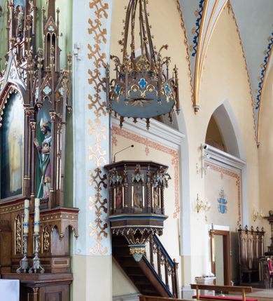 Zdjęcie nr 3: Przyścienną, wiszącą ambonę usytuowano przy łuku tęczowym na zachodniej ścianie prezbiterium. Ambona składa się z baldachimu, zredukowanego zaplecka, kosza oraz prowadzących do niej przyściennych schodów. Wykonana jest z drewna dębowego, bejcowanego i lakierowanego. Partie ornamentalne zostały wyzłocone lub też polichromowane na czerwono i niebiesko. Kosz uzyskał kształt ośmioboczny. Wieńczy go parapet w formie profilowanego gzymsu koronującego. Kolejne pięć boków kosza flankują spiralne kolumienki o liściastych kapitelach ustawionych na wysokich cokolikach, podtrzymujących imposty z pinaklami. Pole każdego boku ozdobiono maswerkowym baldachimem zwieńczonym wygiętą do przodu sterczyną zakończoną kwiatonem. Baldachimy wspierają smukłe kolumienki o liściastych kapitelach ustawione na wysokich cokolikach. Cokoliki kolumienek narożnych i tych podtrzymujących baldachimy wspierają się na łamanym cokole obiegającym cały kosz. Cokół dekorowany od spodu arkaturą złoconą i polichromowaną na czerwono i niebiesko oddziela górną część ambony od jej spodu. Spód ambony utworzono z spływów koncentrujących się na środku na ośmiobocznej, profilowanej opasce dekorowanej od góry stylizowanym motywem lilii heraldycznej, a od dołu stylizowanymi, złoconymi liśćmi winnej latorośli otaczającej dużą kiść winnego grona. Pod baldachimami na czterech bokach umieszczono wykonane w głębokim reliefie przedstawienia stojących ewangelistów. Piąty bok ambony, przylegający do schodów, zdobi zredukowany baldachim pod którym umieszczono złocony i srebrzony kwiat słonecznika. Każdy z ewangelistów stoi na niewielkim postumencie, ustawionym na głównym cokole. Na każdym postumencie widnieję imię przedstawionego ewangelisty. Na południowym boku kosza umieszczono św. Jan. Ukazany został frontalnie z głową ujętą w trzech czwartych w lewo. Ma on długie, falowane, brązowe włosy spływające na ramiona, głowę pochyloną w prawo, wzrok skierowany ku górze. W lewej ręce trzyma księgę, a w prawej, przyciśniętej do piersi pióro. Ubrany jest w długą zieloną tunikę, nakrytą czerwonym płaszczem. Po lewej stronie, u jego stóp widnieje czarny orzeł. Na kolejnym boku znajduje się św. Marek. Wyobrażono go frontalnie z prawą ręką w geście błogosławieństwa, natomiast w lewej trzyma rotulus. Ma on krótkie, brązowe włosy oraz brodę. Ubrany jest w niebieską tunikę odsłaniającą fragmenty bosych stóp i żółty płaszcz. Po lewej stronie przy jego nogach znajduje się lew. Na następnym boku ukazano frontalnie stojącego św. Mateusza. Ewangelista w lewej ręce przyciśniętej do piersi trzyma rotulus, a prawą opuszczoną wzdłóż tułowia wyciąga do przodu. Ma długie, brązowe, falowane włosy spływające na kark oraz brodę. Ubrany jest w blado różową tunikę i żółty płaszcz. Przy jego lewej stopie znajduje się uskrzydlona anielska główka. Dalej znajduje się także przedstawiony frontalnie św. Łukasz. Równocześnie prawą i lewą ręką podtrzymuje zamknietą księgę. Ma on łysiejące czoło i głowę otoczoną siwiejącymi włosami i brodą. Ewangelista ma na sobie brązową tunikę u spodu odsłaniającą fragmenty bosych stóp oraz bladoniebieski płaszcz przerzucony przez prawe ramię. U jego stóp, po lewej stronie, znajduje się wół. Na ambonę prowadzą ulokowane przy ścianie schody o dziesięciu stopniach. Ich balustradę artykułują kolumienki o liściastych kapitelach flankujące ażurowe, ostrołukowe arkady wypełnione u góry złoconym maswerkiem. Zaplecek ambony został zredukowany do szerokiej listwy dekorowanej stylizowaną ostrołukową arkadką. W jej górnej części wyobrażono dwie tablice Dekalogu z numerami przykazań: na jednej: I, II, III, IV, V; na drugiej: VI, VII, VIII, IX, X. Ponad amboną zawieszono wieloboczny baldachim zwieńczony ażurową wiązką spiętych razem pierścieniem zdobionych czołgankami prętów, tworzących dekoracyjny kwiaton wygięty do przodu. Boki baldachimu ozdobiono wycinanymi w desce maswerkami flankowanymi pinaklami, zwieńczonymi powyginanymi sterczynami. Niebieskie podniebie baldachimu zdobi srebrzone wyobrażenie gołębicy Ducha Świętego na tle złocistych promieni. 