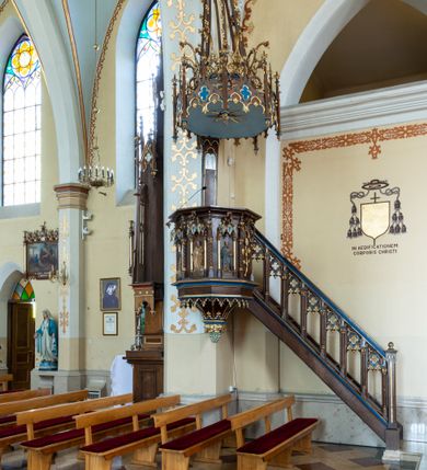 Zdjęcie nr 2: Przyścienną, wiszącą ambonę usytuowano przy łuku tęczowym na zachodniej ścianie prezbiterium. Ambona składa się z baldachimu, zredukowanego zaplecka, kosza oraz prowadzących do niej przyściennych schodów. Wykonana jest z drewna dębowego, bejcowanego i lakierowanego. Partie ornamentalne zostały wyzłocone lub też polichromowane na czerwono i niebiesko. Kosz uzyskał kształt ośmioboczny. Wieńczy go parapet w formie profilowanego gzymsu koronującego. Kolejne pięć boków kosza flankują spiralne kolumienki o liściastych kapitelach ustawionych na wysokich cokolikach, podtrzymujących imposty z pinaklami. Pole każdego boku ozdobiono maswerkowym baldachimem zwieńczonym wygiętą do przodu sterczyną zakończoną kwiatonem. Baldachimy wspierają smukłe kolumienki o liściastych kapitelach ustawione na wysokich cokolikach. Cokoliki kolumienek narożnych i tych podtrzymujących baldachimy wspierają się na łamanym cokole obiegającym cały kosz. Cokół dekorowany od spodu arkaturą złoconą i polichromowaną na czerwono i niebiesko oddziela górną część ambony od jej spodu. Spód ambony utworzono z spływów koncentrujących się na środku na ośmiobocznej, profilowanej opasce dekorowanej od góry stylizowanym motywem lilii heraldycznej, a od dołu stylizowanymi, złoconymi liśćmi winnej latorośli otaczającej dużą kiść winnego grona. Pod baldachimami na czterech bokach umieszczono wykonane w głębokim reliefie przedstawienia stojących ewangelistów. Piąty bok ambony, przylegający do schodów, zdobi zredukowany baldachim pod którym umieszczono złocony i srebrzony kwiat słonecznika. Każdy z ewangelistów stoi na niewielkim postumencie, ustawionym na głównym cokole. Na każdym postumencie widnieję imię przedstawionego ewangelisty. Na południowym boku kosza umieszczono św. Jan. Ukazany został frontalnie z głową ujętą w trzech czwartych w lewo. Ma on długie, falowane, brązowe włosy spływające na ramiona, głowę pochyloną w prawo, wzrok skierowany ku górze. W lewej ręce trzyma księgę, a w prawej, przyciśniętej do piersi pióro. Ubrany jest w długą zieloną tunikę, nakrytą czerwonym płaszczem. Po lewej stronie, u jego stóp widnieje czarny orzeł. Na kolejnym boku znajduje się św. Marek. Wyobrażono go frontalnie z prawą ręką w geście błogosławieństwa, natomiast w lewej trzyma rotulus. Ma on krótkie, brązowe włosy oraz brodę. Ubrany jest w niebieską tunikę odsłaniającą fragmenty bosych stóp i żółty płaszcz. Po lewej stronie przy jego nogach znajduje się lew. Na następnym boku ukazano frontalnie stojącego św. Mateusza. Ewangelista w lewej ręce przyciśniętej do piersi trzyma rotulus, a prawą opuszczoną wzdłóż tułowia wyciąga do przodu. Ma długie, brązowe, falowane włosy spływające na kark oraz brodę. Ubrany jest w blado różową tunikę i żółty płaszcz. Przy jego lewej stopie znajduje się uskrzydlona anielska główka. Dalej znajduje się także przedstawiony frontalnie św. Łukasz. Równocześnie prawą i lewą ręką podtrzymuje zamknietą księgę. Ma on łysiejące czoło i głowę otoczoną siwiejącymi włosami i brodą. Ewangelista ma na sobie brązową tunikę u spodu odsłaniającą fragmenty bosych stóp oraz bladoniebieski płaszcz przerzucony przez prawe ramię. U jego stóp, po lewej stronie, znajduje się wół. Na ambonę prowadzą ulokowane przy ścianie schody o dziesięciu stopniach. Ich balustradę artykułują kolumienki o liściastych kapitelach flankujące ażurowe, ostrołukowe arkady wypełnione u góry złoconym maswerkiem. Zaplecek ambony został zredukowany do szerokiej listwy dekorowanej stylizowaną ostrołukową arkadką. W jej górnej części wyobrażono dwie tablice Dekalogu z numerami przykazań: na jednej: I, II, III, IV, V; na drugiej: VI, VII, VIII, IX, X. Ponad amboną zawieszono wieloboczny baldachim zwieńczony ażurową wiązką spiętych razem pierścieniem zdobionych czołgankami prętów, tworzących dekoracyjny kwiaton wygięty do przodu. Boki baldachimu ozdobiono wycinanymi w desce maswerkami flankowanymi pinaklami, zwieńczonymi powyginanymi sterczynami. Niebieskie podniebie baldachimu zdobi srebrzone wyobrażenie gołębicy Ducha Świętego na tle złocistych promieni. 