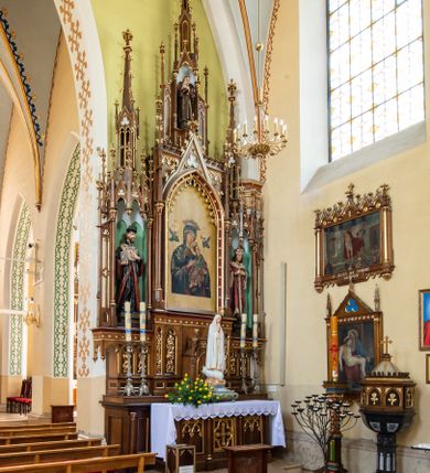 Zdjęcie nr 4: Neogotycki, przyścienny, jednokondygnacyjny ołtarz Świętej Rodziny uzyskał architektoniczną strukturę ze zwieńczeniem. Kolorystyka ołtarza utrzymana została w tonacji ciemnego, bejcowanego drewna. Detale, ornamenty zostały wyzłocone, natomiast wypełnienia maswerków i ram wimperg są białe. Z kolei pole ramy głównego obrazu wykonano jako czerwone. Retabulum ustawiono na podwójnych gradusach zaokrąglonych od przodu. Stipes ołtarzowy uzyskał kształt prostopadłościanu z trzema czworobocznymi płycinami od frontu, ujętymi złoconymi i profilowanymi obramieniami. Płyciny rozdzielają półkolumienki na wysokich cokolikach. Dwie skrajne płyciny stipesu wypełniono u góry maswerkiem, natomiast w środkowej znalazł się równoramienny krzyż wpisany w czteroliść. Stipes podtrzymuje drewnianą mensę z portatylem. Ołtarz przylega do wysokiego, prostopadłościennego cokołu flankowanego po bokach kolumienkami, na którym wspiera się cała nastawa. Widoczne jego partie zdobią prostokątne płyciny w obramieniach. Na cokole wspiera się predella podzielona płytkimi, prostokątnymi płycinami zakończonymi ostrołukowymi arkadkami z noskami, wypełnionymi płaskorzeźbionym i złoconym ornamentem roślinnym. Pośrodku, w przerwie między płycinami predelli, usytuowano prostopadłościenne tabernakulum w formie kapliczki, zwieńczone łukiem siodłowym, flankowane dwoma pinaklami. Ostrołukowe drzwiczki tabernakulum zdobi skrzyżowana z krzyżem kotwica oraz wyobrażenie Boskiego Serca Pana Jezusa. Poniżej widoczna jest girlanda różana. Nad drzwiczkami umieszczono stylizowany ornament roślinny. Predella wspiera trójosiową kondygnację ze zwieńczeniem. Na osi środkowej usytuowano płytką, prostokątną wnękę zakończoną ostrołukiem z arkaturą. Flankują ją smukłe kolumienki na wysokich cokolikach wspierające wysokie pinakle. Ponad wnęką widoczna jest wimperga z maswerkiem dekorowana czołgankami i kwiatonem. We wnęce umieszczono pełnoplastyczne rzeźby Matki Boskiej i św. Józefa, pomiędzy którymi stoi mały Jezus, a na zasuwie obraz Matki Boskiej Nieustającej Pomocy. W osiach skrajnych, pod gotyckimi baldachimami z wimpergami, rozdzielonymi pinaklami, ustawiono w niszach na postumentach figury św. Jana Kantego i św. Kazimierza Królewicza. Święci są podpisani na postumentach. Konchy nisz są jasnoniebieskie natomiast ich ścianki zielone. Boki baldachimów udekorowano małymi wimpergami rozdzielonymi smukłymi pinaklami z kwiatonami i czołgankami. Baldachimy przechodzą w smukłe, ażurowe fiale dekorowane pinaklami i czołgankami oraz kwiatonami. Zwieńczenie nastawy na osi środkowej uzyskało kształt otwartej kapliczki z niszą zakończoną smukłą fialą z iglicą, dekorowaną czołgankami i kwiatonem. Po bokach kapliczki znajdują się przypory dekorowane maswerkiem i flankowane pinaklami. W niszy umieszczono figurę św. Antoniego z Padwy, którego podpisano na postumencie. Całość retabulum flankują uszaki w formie ażurowych przypór, wypełnionych maswerkami i zdobionych parą pinakli.
