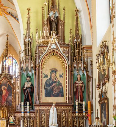 Zdjęcie nr 3: Neogotycki, przyścienny, jednokondygnacyjny ołtarz Świętej Rodziny uzyskał architektoniczną strukturę ze zwieńczeniem. Kolorystyka ołtarza utrzymana została w tonacji ciemnego, bejcowanego drewna. Detale, ornamenty zostały wyzłocone, natomiast wypełnienia maswerków i ram wimperg są białe. Z kolei pole ramy głównego obrazu wykonano jako czerwone. Retabulum ustawiono na podwójnych gradusach zaokrąglonych od przodu. Stipes ołtarzowy uzyskał kształt prostopadłościanu z trzema czworobocznymi płycinami od frontu, ujętymi złoconymi i profilowanymi obramieniami. Płyciny rozdzielają półkolumienki na wysokich cokolikach. Dwie skrajne płyciny stipesu wypełniono u góry maswerkiem, natomiast w środkowej znalazł się równoramienny krzyż wpisany w czteroliść. Stipes podtrzymuje drewnianą mensę z portatylem. Ołtarz przylega do wysokiego, prostopadłościennego cokołu flankowanego po bokach kolumienkami, na którym wspiera się cała nastawa. Widoczne jego partie zdobią prostokątne płyciny w obramieniach. Na cokole wspiera się predella podzielona płytkimi, prostokątnymi płycinami zakończonymi ostrołukowymi arkadkami z noskami, wypełnionymi płaskorzeźbionym i złoconym ornamentem roślinnym. Pośrodku, w przerwie między płycinami predelli, usytuowano prostopadłościenne tabernakulum w formie kapliczki, zwieńczone łukiem siodłowym, flankowane dwoma pinaklami. Ostrołukowe drzwiczki tabernakulum zdobi skrzyżowana z krzyżem kotwica oraz wyobrażenie Boskiego Serca Pana Jezusa. Poniżej widoczna jest girlanda różana. Nad drzwiczkami umieszczono stylizowany ornament roślinny. Predella wspiera trójosiową kondygnację ze zwieńczeniem. Na osi środkowej usytuowano płytką, prostokątną wnękę zakończoną ostrołukiem z arkaturą. Flankują ją smukłe kolumienki na wysokich cokolikach wspierające wysokie pinakle. Ponad wnęką widoczna jest wimperga z maswerkiem dekorowana czołgankami i kwiatonem. We wnęce umieszczono pełnoplastyczne rzeźby Matki Boskiej i św. Józefa, pomiędzy którymi stoi mały Jezus, a na zasuwie obraz Matki Boskiej Nieustającej Pomocy. W osiach skrajnych, pod gotyckimi baldachimami z wimpergami, rozdzielonymi pinaklami, ustawiono w niszach na postumentach figury św. Jana Kantego i św. Kazimierza Królewicza. Święci są podpisani na postumentach. Konchy nisz są jasnoniebieskie natomiast ich ścianki zielone. Boki baldachimów udekorowano małymi wimpergami rozdzielonymi smukłymi pinaklami z kwiatonami i czołgankami. Baldachimy przechodzą w smukłe, ażurowe fiale dekorowane pinaklami i czołgankami oraz kwiatonami. Zwieńczenie nastawy na osi środkowej uzyskało kształt otwartej kapliczki z niszą zakończoną smukłą fialą z iglicą, dekorowaną czołgankami i kwiatonem. Po bokach kapliczki znajdują się przypory dekorowane maswerkiem i flankowane pinaklami. W niszy umieszczono figurę św. Antoniego z Padwy, którego podpisano na postumencie. Całość retabulum flankują uszaki w formie ażurowych przypór, wypełnionych maswerkami i zdobionych parą pinakli.
