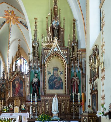 Zdjęcie nr 1: Neogotycki, przyścienny, jednokondygnacyjny ołtarz Świętej Rodziny uzyskał architektoniczną strukturę ze zwieńczeniem. Kolorystyka ołtarza utrzymana została w tonacji ciemnego, bejcowanego drewna. Detale, ornamenty zostały wyzłocone, natomiast wypełnienia maswerków i ram wimperg są białe. Z kolei pole ramy głównego obrazu wykonano jako czerwone. Retabulum ustawiono na podwójnych gradusach zaokrąglonych od przodu. Stipes ołtarzowy uzyskał kształt prostopadłościanu z trzema czworobocznymi płycinami od frontu, ujętymi złoconymi i profilowanymi obramieniami. Płyciny rozdzielają półkolumienki na wysokich cokolikach. Dwie skrajne płyciny stipesu wypełniono u góry maswerkiem, natomiast w środkowej znalazł się równoramienny krzyż wpisany w czteroliść. Stipes podtrzymuje drewnianą mensę z portatylem. Ołtarz przylega do wysokiego, prostopadłościennego cokołu flankowanego po bokach kolumienkami, na którym wspiera się cała nastawa. Widoczne jego partie zdobią prostokątne płyciny w obramieniach. Na cokole wspiera się predella podzielona płytkimi, prostokątnymi płycinami zakończonymi ostrołukowymi arkadkami z noskami, wypełnionymi płaskorzeźbionym i złoconym ornamentem roślinnym. Pośrodku, w przerwie między płycinami predelli, usytuowano prostopadłościenne tabernakulum w formie kapliczki, zwieńczone łukiem siodłowym, flankowane dwoma pinaklami. Ostrołukowe drzwiczki tabernakulum zdobi skrzyżowana z krzyżem kotwica oraz wyobrażenie Boskiego Serca Pana Jezusa. Poniżej widoczna jest girlanda różana. Nad drzwiczkami umieszczono stylizowany ornament roślinny. Predella wspiera trójosiową kondygnację ze zwieńczeniem. Na osi środkowej usytuowano płytką, prostokątną wnękę zakończoną ostrołukiem z arkaturą. Flankują ją smukłe kolumienki na wysokich cokolikach wspierające wysokie pinakle. Ponad wnęką widoczna jest wimperga z maswerkiem dekorowana czołgankami i kwiatonem. We wnęce umieszczono pełnoplastyczne rzeźby Matki Boskiej i św. Józefa, pomiędzy którymi stoi mały Jezus, a na zasuwie obraz Matki Boskiej Nieustającej Pomocy. W osiach skrajnych, pod gotyckimi baldachimami z wimpergami, rozdzielonymi pinaklami, ustawiono w niszach na postumentach figury św. Jana Kantego i św. Kazimierza Królewicza. Święci są podpisani na postumentach. Konchy nisz są jasnoniebieskie natomiast ich ścianki zielone. Boki baldachimów udekorowano małymi wimpergami rozdzielonymi smukłymi pinaklami z kwiatonami i czołgankami. Baldachimy przechodzą w smukłe, ażurowe fiale dekorowane pinaklami i czołgankami oraz kwiatonami. Zwieńczenie nastawy na osi środkowej uzyskało kształt otwartej kapliczki z niszą zakończoną smukłą fialą z iglicą, dekorowaną czołgankami i kwiatonem. Po bokach kapliczki znajdują się przypory dekorowane maswerkiem i flankowane pinaklami. W niszy umieszczono figurę św. Antoniego z Padwy, którego podpisano na postumencie. Całość retabulum flankują uszaki w formie ażurowych przypór, wypełnionych maswerkami i zdobionych parą pinakli.
