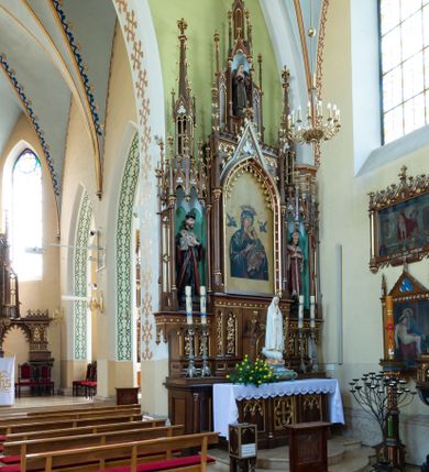 Zdjęcie nr 2: Neogotycki, przyścienny, jednokondygnacyjny ołtarz Świętej Rodziny uzyskał architektoniczną strukturę ze zwieńczeniem. Kolorystyka ołtarza utrzymana została w tonacji ciemnego, bejcowanego drewna. Detale, ornamenty zostały wyzłocone, natomiast wypełnienia maswerków i ram wimperg są białe. Z kolei pole ramy głównego obrazu wykonano jako czerwone. Retabulum ustawiono na podwójnych gradusach zaokrąglonych od przodu. Stipes ołtarzowy uzyskał kształt prostopadłościanu z trzema czworobocznymi płycinami od frontu, ujętymi złoconymi i profilowanymi obramieniami. Płyciny rozdzielają półkolumienki na wysokich cokolikach. Dwie skrajne płyciny stipesu wypełniono u góry maswerkiem, natomiast w środkowej znalazł się równoramienny krzyż wpisany w czteroliść. Stipes podtrzymuje drewnianą mensę z portatylem. Ołtarz przylega do wysokiego, prostopadłościennego cokołu flankowanego po bokach kolumienkami, na którym wspiera się cała nastawa. Widoczne jego partie zdobią prostokątne płyciny w obramieniach. Na cokole wspiera się predella podzielona płytkimi, prostokątnymi płycinami zakończonymi ostrołukowymi arkadkami z noskami, wypełnionymi płaskorzeźbionym i złoconym ornamentem roślinnym. Pośrodku, w przerwie między płycinami predelli, usytuowano prostopadłościenne tabernakulum w formie kapliczki, zwieńczone łukiem siodłowym, flankowane dwoma pinaklami. Ostrołukowe drzwiczki tabernakulum zdobi skrzyżowana z krzyżem kotwica oraz wyobrażenie Boskiego Serca Pana Jezusa. Poniżej widoczna jest girlanda różana. Nad drzwiczkami umieszczono stylizowany ornament roślinny. Predella wspiera trójosiową kondygnację ze zwieńczeniem. Na osi środkowej usytuowano płytką, prostokątną wnękę zakończoną ostrołukiem z arkaturą. Flankują ją smukłe kolumienki na wysokich cokolikach wspierające wysokie pinakle. Ponad wnęką widoczna jest wimperga z maswerkiem dekorowana czołgankami i kwiatonem. We wnęce umieszczono pełnoplastyczne rzeźby Matki Boskiej i św. Józefa, pomiędzy którymi stoi mały Jezus, a na zasuwie obraz Matki Boskiej Nieustającej Pomocy. W osiach skrajnych, pod gotyckimi baldachimami z wimpergami, rozdzielonymi pinaklami, ustawiono w niszach na postumentach figury św. Jana Kantego i św. Kazimierza Królewicza. Święci są podpisani na postumentach. Konchy nisz są jasnoniebieskie natomiast ich ścianki zielone. Boki baldachimów udekorowano małymi wimpergami rozdzielonymi smukłymi pinaklami z kwiatonami i czołgankami. Baldachimy przechodzą w smukłe, ażurowe fiale dekorowane pinaklami i czołgankami oraz kwiatonami. Zwieńczenie nastawy na osi środkowej uzyskało kształt otwartej kapliczki z niszą zakończoną smukłą fialą z iglicą, dekorowaną czołgankami i kwiatonem. Po bokach kapliczki znajdują się przypory dekorowane maswerkiem i flankowane pinaklami. W niszy umieszczono figurę św. Antoniego z Padwy, którego podpisano na postumencie. Całość retabulum flankują uszaki w formie ażurowych przypór, wypełnionych maswerkami i zdobionych parą pinakli.
