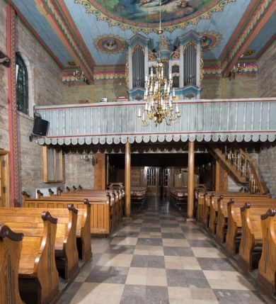 Zdjęcie nr 11: Kościół, otoczony starodrzewiem, położony jest na niewielkim wzniesieniu ponad stawami w centrum wsi. Jest to budowla drewniano-murowana, orientowana. Nawa została wzniesiona w 1518 roku na zrąb na rzucie zbliżonym do kwadratu, nakryta płaskim stropem. Od zachodu do nawy przylega wzniesiona w tym samym roku wieża o konstrukcji słupowej, pierwotnie kwadratowa o pochyłych ścianach, w 1782 roku poszerzona przez obudowanie drugą konstrukcją, prawie pionową. Wieża, w całości szalowana, poprzedzona niewielką kruchtą, w połowie wysokości jest opasana daszkiem pulpitowym. Jej hełm posiada kształt kopuły zwieńczonej ostrosłupem. Od południa do nawy przylega drewniana kruchta z 1924 roku, a od wschodu murowane prezbiterium z 1931 roku. Prezbiterium otwiera się podwójnymi arkadami na dwie kaplice boczne - wzniesioną od południa w 1861 roku z kryptą grobową Duninów oraz kaplicę przylegającą od północy zbudowaną w 1931 roku. Jego przestrzeń nakryto sklepieniem kolebkowym na gurtach, zaś dwuprzęsłowe kaplice sklepieniem krzyżowym. Za prezbiterium wznosi się murowana, piętrowa zakrystia z XVII wieku przykryta sklepieniem nieckowym z lunetami i małym zwierciadłem, zdobionym monogramem maryjnym. Jednokalenicowy dach nawy i prezbiterium pokryty jest dachówką. Nad końcu dachu nawy od wschodu nadwieszono wieżyczkę na sygnaturkę z latarnią i cebulastą banią, zwieńczoną krzyżem. Zakrystia nakryta została namiotowym dachem polskim, także pokrytym dachówką. Na zewnętrznych ścianach części murowanej umieszczono kilka epitafiów przeniesionych z wnętrza kościoła. Wewnątrz prezbiterium na ścianie wschodniej, nad wejściem do zakrystii, zawieszono manierystyczny, dwukondygnacyjny ołtarz główny z XVII wieku z uszakami zdobionymi herbami Starykoń i Jelita. Przy ścianie północnej, w miejscu połączenia nawy drewnianej z murowanym prezbiterium, usytuowano w 1931 roku ambonę oraz kamienną chrzcielnicę z 1658 roku. Belkę tęczową zdobi osiemnastowieczna grupa Ukrzyżowania. W kaplicach bocznych ustawiono dwa neobarokowe ołtarze boczne.  