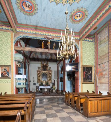 Zdjęcie nr 6: Kościół, otoczony starodrzewiem, położony jest na niewielkim wzniesieniu ponad stawami w centrum wsi. Jest to budowla drewniano-murowana, orientowana. Nawa została wzniesiona w 1518 roku na zrąb na rzucie zbliżonym do kwadratu, nakryta płaskim stropem. Od zachodu do nawy przylega wzniesiona w tym samym roku wieża o konstrukcji słupowej, pierwotnie kwadratowa o pochyłych ścianach, w 1782 roku poszerzona przez obudowanie drugą konstrukcją, prawie pionową. Wieża, w całości szalowana, poprzedzona niewielką kruchtą, w połowie wysokości jest opasana daszkiem pulpitowym. Jej hełm posiada kształt kopuły zwieńczonej ostrosłupem. Od południa do nawy przylega drewniana kruchta z 1924 roku, a od wschodu murowane prezbiterium z 1931 roku. Prezbiterium otwiera się podwójnymi arkadami na dwie kaplice boczne - wzniesioną od południa w 1861 roku z kryptą grobową Duninów oraz kaplicę przylegającą od północy zbudowaną w 1931 roku. Jego przestrzeń nakryto sklepieniem kolebkowym na gurtach, zaś dwuprzęsłowe kaplice sklepieniem krzyżowym. Za prezbiterium wznosi się murowana, piętrowa zakrystia z XVII wieku przykryta sklepieniem nieckowym z lunetami i małym zwierciadłem, zdobionym monogramem maryjnym. Jednokalenicowy dach nawy i prezbiterium pokryty jest dachówką. Nad końcu dachu nawy od wschodu nadwieszono wieżyczkę na sygnaturkę z latarnią i cebulastą banią, zwieńczoną krzyżem. Zakrystia nakryta została namiotowym dachem polskim, także pokrytym dachówką. Na zewnętrznych ścianach części murowanej umieszczono kilka epitafiów przeniesionych z wnętrza kościoła. Wewnątrz prezbiterium na ścianie wschodniej, nad wejściem do zakrystii, zawieszono manierystyczny, dwukondygnacyjny ołtarz główny z XVII wieku z uszakami zdobionymi herbami Starykoń i Jelita. Przy ścianie północnej, w miejscu połączenia nawy drewnianej z murowanym prezbiterium, usytuowano w 1931 roku ambonę oraz kamienną chrzcielnicę z 1658 roku. Belkę tęczową zdobi osiemnastowieczna grupa Ukrzyżowania. W kaplicach bocznych ustawiono dwa neobarokowe ołtarze boczne.  