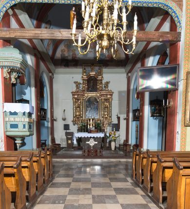 Zdjęcie nr 9: Kościół, otoczony starodrzewiem, położony jest na niewielkim wzniesieniu ponad stawami w centrum wsi. Jest to budowla drewniano-murowana, orientowana. Nawa została wzniesiona w 1518 roku na zrąb na rzucie zbliżonym do kwadratu, nakryta płaskim stropem. Od zachodu do nawy przylega wzniesiona w tym samym roku wieża o konstrukcji słupowej, pierwotnie kwadratowa o pochyłych ścianach, w 1782 roku poszerzona przez obudowanie drugą konstrukcją, prawie pionową. Wieża, w całości szalowana, poprzedzona niewielką kruchtą, w połowie wysokości jest opasana daszkiem pulpitowym. Jej hełm posiada kształt kopuły zwieńczonej ostrosłupem. Od południa do nawy przylega drewniana kruchta z 1924 roku, a od wschodu murowane prezbiterium z 1931 roku. Prezbiterium otwiera się podwójnymi arkadami na dwie kaplice boczne - wzniesioną od południa w 1861 roku z kryptą grobową Duninów oraz kaplicę przylegającą od północy zbudowaną w 1931 roku. Jego przestrzeń nakryto sklepieniem kolebkowym na gurtach, zaś dwuprzęsłowe kaplice sklepieniem krzyżowym. Za prezbiterium wznosi się murowana, piętrowa zakrystia z XVII wieku przykryta sklepieniem nieckowym z lunetami i małym zwierciadłem, zdobionym monogramem maryjnym. Jednokalenicowy dach nawy i prezbiterium pokryty jest dachówką. Nad końcu dachu nawy od wschodu nadwieszono wieżyczkę na sygnaturkę z latarnią i cebulastą banią, zwieńczoną krzyżem. Zakrystia nakryta została namiotowym dachem polskim, także pokrytym dachówką. Na zewnętrznych ścianach części murowanej umieszczono kilka epitafiów przeniesionych z wnętrza kościoła. Wewnątrz prezbiterium na ścianie wschodniej, nad wejściem do zakrystii, zawieszono manierystyczny, dwukondygnacyjny ołtarz główny z XVII wieku z uszakami zdobionymi herbami Starykoń i Jelita. Przy ścianie północnej, w miejscu połączenia nawy drewnianej z murowanym prezbiterium, usytuowano w 1931 roku ambonę oraz kamienną chrzcielnicę z 1658 roku. Belkę tęczową zdobi osiemnastowieczna grupa Ukrzyżowania. W kaplicach bocznych ustawiono dwa neobarokowe ołtarze boczne.  