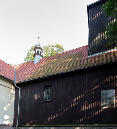 Zdjęcie nr 19: Kościół, otoczony starodrzewiem, położony jest na niewielkim wzniesieniu ponad stawami w centrum wsi. Jest to budowla drewniano-murowana, orientowana. Nawa została wzniesiona w 1518 roku na zrąb na rzucie zbliżonym do kwadratu, nakryta płaskim stropem. Od zachodu do nawy przylega wzniesiona w tym samym roku wieża o konstrukcji słupowej, pierwotnie kwadratowa o pochyłych ścianach, w 1782 roku poszerzona przez obudowanie drugą konstrukcją, prawie pionową. Wieża, w całości szalowana, poprzedzona niewielką kruchtą, w połowie wysokości jest opasana daszkiem pulpitowym. Jej hełm posiada kształt kopuły zwieńczonej ostrosłupem. Od południa do nawy przylega drewniana kruchta z 1924 roku, a od wschodu murowane prezbiterium z 1931 roku. Prezbiterium otwiera się podwójnymi arkadami na dwie kaplice boczne - wzniesioną od południa w 1861 roku z kryptą grobową Duninów oraz kaplicę przylegającą od północy zbudowaną w 1931 roku. Jego przestrzeń nakryto sklepieniem kolebkowym na gurtach, zaś dwuprzęsłowe kaplice sklepieniem krzyżowym. Za prezbiterium wznosi się murowana, piętrowa zakrystia z XVII wieku przykryta sklepieniem nieckowym z lunetami i małym zwierciadłem, zdobionym monogramem maryjnym. Jednokalenicowy dach nawy i prezbiterium pokryty jest dachówką. Nad końcu dachu nawy od wschodu nadwieszono wieżyczkę na sygnaturkę z latarnią i cebulastą banią, zwieńczoną krzyżem. Zakrystia nakryta została namiotowym dachem polskim, także pokrytym dachówką. Na zewnętrznych ścianach części murowanej umieszczono kilka epitafiów przeniesionych z wnętrza kościoła. Wewnątrz prezbiterium na ścianie wschodniej, nad wejściem do zakrystii, zawieszono manierystyczny, dwukondygnacyjny ołtarz główny z XVII wieku z uszakami zdobionymi herbami Starykoń i Jelita. Przy ścianie północnej, w miejscu połączenia nawy drewnianej z murowanym prezbiterium, usytuowano w 1931 roku ambonę oraz kamienną chrzcielnicę z 1658 roku. Belkę tęczową zdobi osiemnastowieczna grupa Ukrzyżowania. W kaplicach bocznych ustawiono dwa neobarokowe ołtarze boczne.  