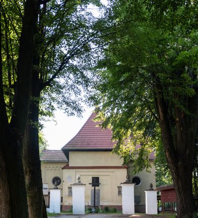 Zdjęcie nr 21: Kościół, otoczony starodrzewiem, położony jest na niewielkim wzniesieniu ponad stawami w centrum wsi. Jest to budowla drewniano-murowana, orientowana. Nawa została wzniesiona w 1518 roku na zrąb na rzucie zbliżonym do kwadratu, nakryta płaskim stropem. Od zachodu do nawy przylega wzniesiona w tym samym roku wieża o konstrukcji słupowej, pierwotnie kwadratowa o pochyłych ścianach, w 1782 roku poszerzona przez obudowanie drugą konstrukcją, prawie pionową. Wieża, w całości szalowana, poprzedzona niewielką kruchtą, w połowie wysokości jest opasana daszkiem pulpitowym. Jej hełm posiada kształt kopuły zwieńczonej ostrosłupem. Od południa do nawy przylega drewniana kruchta z 1924 roku, a od wschodu murowane prezbiterium z 1931 roku. Prezbiterium otwiera się podwójnymi arkadami na dwie kaplice boczne - wzniesioną od południa w 1861 roku z kryptą grobową Duninów oraz kaplicę przylegającą od północy zbudowaną w 1931 roku. Jego przestrzeń nakryto sklepieniem kolebkowym na gurtach, zaś dwuprzęsłowe kaplice sklepieniem krzyżowym. Za prezbiterium wznosi się murowana, piętrowa zakrystia z XVII wieku przykryta sklepieniem nieckowym z lunetami i małym zwierciadłem, zdobionym monogramem maryjnym. Jednokalenicowy dach nawy i prezbiterium pokryty jest dachówką. Nad końcu dachu nawy od wschodu nadwieszono wieżyczkę na sygnaturkę z latarnią i cebulastą banią, zwieńczoną krzyżem. Zakrystia nakryta została namiotowym dachem polskim, także pokrytym dachówką. Na zewnętrznych ścianach części murowanej umieszczono kilka epitafiów przeniesionych z wnętrza kościoła. Wewnątrz prezbiterium na ścianie wschodniej, nad wejściem do zakrystii, zawieszono manierystyczny, dwukondygnacyjny ołtarz główny z XVII wieku z uszakami zdobionymi herbami Starykoń i Jelita. Przy ścianie północnej, w miejscu połączenia nawy drewnianej z murowanym prezbiterium, usytuowano w 1931 roku ambonę oraz kamienną chrzcielnicę z 1658 roku. Belkę tęczową zdobi osiemnastowieczna grupa Ukrzyżowania. W kaplicach bocznych ustawiono dwa neobarokowe ołtarze boczne.  