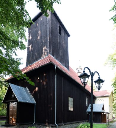Zdjęcie nr 1: Kościół, otoczony starodrzewiem, położony jest na niewielkim wzniesieniu ponad stawami w centrum wsi. Jest to budowla drewniano-murowana, orientowana. Nawa została wzniesiona w 1518 roku na zrąb na rzucie zbliżonym do kwadratu, nakryta płaskim stropem. Od zachodu do nawy przylega wzniesiona w tym samym roku wieża o konstrukcji słupowej, pierwotnie kwadratowa o pochyłych ścianach, w 1782 roku poszerzona przez obudowanie drugą konstrukcją, prawie pionową. Wieża, w całości szalowana, poprzedzona niewielką kruchtą, w połowie wysokości jest opasana daszkiem pulpitowym. Jej hełm posiada kształt kopuły zwieńczonej ostrosłupem. Od południa do nawy przylega drewniana kruchta z 1924 roku, a od wschodu murowane prezbiterium z 1931 roku. Prezbiterium otwiera się podwójnymi arkadami na dwie kaplice boczne - wzniesioną od południa w 1861 roku z kryptą grobową Duninów oraz kaplicę przylegającą od północy zbudowaną w 1931 roku. Jego przestrzeń nakryto sklepieniem kolebkowym na gurtach, zaś dwuprzęsłowe kaplice sklepieniem krzyżowym. Za prezbiterium wznosi się murowana, piętrowa zakrystia z XVII wieku przykryta sklepieniem nieckowym z lunetami i małym zwierciadłem, zdobionym monogramem maryjnym. Jednokalenicowy dach nawy i prezbiterium pokryty jest dachówką. Nad końcu dachu nawy od wschodu nadwieszono wieżyczkę na sygnaturkę z latarnią i cebulastą banią, zwieńczoną krzyżem. Zakrystia nakryta została namiotowym dachem polskim, także pokrytym dachówką. Na zewnętrznych ścianach części murowanej umieszczono kilka epitafiów przeniesionych z wnętrza kościoła. Wewnątrz prezbiterium na ścianie wschodniej, nad wejściem do zakrystii, zawieszono manierystyczny, dwukondygnacyjny ołtarz główny z XVII wieku z uszakami zdobionymi herbami Starykoń i Jelita. Przy ścianie północnej, w miejscu połączenia nawy drewnianej z murowanym prezbiterium, usytuowano w 1931 roku ambonę oraz kamienną chrzcielnicę z 1658 roku. Belkę tęczową zdobi osiemnastowieczna grupa Ukrzyżowania. W kaplicach bocznych ustawiono dwa neobarokowe ołtarze boczne.  
