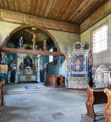 Zdjęcie nr 9: Kościół jest usytuowany ustronnie na stoku wzniesienia, otoczony starodrzewiem i parkanem, ujmującym stary cmentarz z nielicznymi mogiłami i nagrobkami. Orientowaną świątynię w całości wzniesiono z drewna. Zastosowano konstrukcję zrębową na murowanej podwalinie z murowaną kryptą pod prezbiterium. Nawa kościoła jest prawie kwadratowa. Od wchodu przylega do niej węższe prezbiterium zamknięte trójbocznie. Przy jego północnej ścianie wzniesiono bezokienną zakrystię z wejściem od strony wschodniej. Od zachodu z nawą łączy się wieża na rzucie kwadratu wzniesiona na tzw. słup. Ma pochyłe ściany, szalowane w dolnej części deskami, a w górnej podbite gontem. Wieża zakończona jest nadwieszoną izbicą, zwieńczoną ostrosłupowym hełmem gontowym. W połowie wysokości wieżę opasa pulpitowy daszek gontowy łączący się po bokach z dachem nawy. Pod wieżą urządzono obszerną kruchtę. Wysokie i spadziste dachy kościoła pokryte są gontem. Więźba ma konstrukcję storczykową. Ponad wschodnim szczytem nawy usytuowano nadwieszoną wieżyczkę na sygnaturkę o kształcie cebulastym z latarnią zwieńczoną krzyżem. Całość bryły kościoła (z wyjątkiem wieży) obiegają otwarte soboty, w które przed zakrystią od wschodu wbudowano nowy przedsionek. W nawie od południa znajduje się profilowany portal z nadprożem wykrojonym w ośli grzbiet. Umieszczone w nim drzwi zostały zaopatrzone w stare okucia i antabę. Wnętrze nawy i prezbiterium nakryto płaskimi stropami z pozostałościami późnorenesansowych malowideł z motywem kasetonów z rozetami. Łuk tęczowy jest ostry. Spływa na profilowaną belkę, która jest wsparta na kroksztynach. Na belce tęczowej został ustawiony krucyfiks oraz figury Matki Boskiej i św. Jana Apostoła. Chór muzyczny z dekorowanym malarsko parapetem został wsparty na dwóch słupach. W świątyni znajduje się rokokowy ołtarz główny z obrazem ze sceną Zwiastowania NMP. Dwa ołtarze boczne ustawione zostały przy łuku tęczowym. Prawy ołtarz boczny św. Karola Boromeusza pochodzi z drugiej połowy XVII wieku. Na to miejsce trafił on w połowie XX wieku z kaplicy pałacu w Osieku. Lewy ołtarz boczny jest późnobarokowy z około 1700 roku. W jego centrum umieszczono obraz Matki Boskiej Piekarskiej. Na północnej ścianie nad wejściem do zakrystii zawieszono epitafium Mikołaja i Beaty z Mirowa Porębskich z 1611 roku. Wcześniej epitafium pełniło rolę retabulum w miejscu stojącego dziś ołtarza św. Karola Boromeusza. W prezbiterium przy łuku tęczowym zawieszono rokokową ambonę w kształcie łodzi, na którą wejście prowadzi z zakrystii. Przy wejściu do nawy znajduje się kamienna chrzcielnica z początku XVI wieku, ozdobiona herbem Fogelweder, z drewnianą, późnobarokową nakrywą. W zakrystii zachował się także rokokowy drewniany lawetarz z drugiej połowy XVIII wieku.     