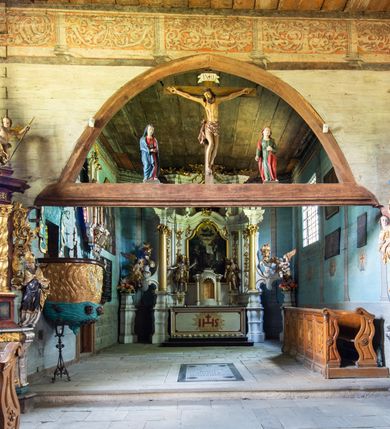 Zdjęcie nr 7: Kościół jest usytuowany ustronnie na stoku wzniesienia, otoczony starodrzewiem i parkanem, ujmującym stary cmentarz z nielicznymi mogiłami i nagrobkami. Orientowaną świątynię w całości wzniesiono z drewna. Zastosowano konstrukcję zrębową na murowanej podwalinie z murowaną kryptą pod prezbiterium. Nawa kościoła jest prawie kwadratowa. Od wchodu przylega do niej węższe prezbiterium zamknięte trójbocznie. Przy jego północnej ścianie wzniesiono bezokienną zakrystię z wejściem od strony wschodniej. Od zachodu z nawą łączy się wieża na rzucie kwadratu wzniesiona na tzw. słup. Ma pochyłe ściany, szalowane w dolnej części deskami, a w górnej podbite gontem. Wieża zakończona jest nadwieszoną izbicą, zwieńczoną ostrosłupowym hełmem gontowym. W połowie wysokości wieżę opasa pulpitowy daszek gontowy łączący się po bokach z dachem nawy. Pod wieżą urządzono obszerną kruchtę. Wysokie i spadziste dachy kościoła pokryte są gontem. Więźba ma konstrukcję storczykową. Ponad wschodnim szczytem nawy usytuowano nadwieszoną wieżyczkę na sygnaturkę o kształcie cebulastym z latarnią zwieńczoną krzyżem. Całość bryły kościoła (z wyjątkiem wieży) obiegają otwarte soboty, w które przed zakrystią od wschodu wbudowano nowy przedsionek. W nawie od południa znajduje się profilowany portal z nadprożem wykrojonym w ośli grzbiet. Umieszczone w nim drzwi zostały zaopatrzone w stare okucia i antabę. Wnętrze nawy i prezbiterium nakryto płaskimi stropami z pozostałościami późnorenesansowych malowideł z motywem kasetonów z rozetami. Łuk tęczowy jest ostry. Spływa na profilowaną belkę, która jest wsparta na kroksztynach. Na belce tęczowej został ustawiony krucyfiks oraz figury Matki Boskiej i św. Jana Apostoła. Chór muzyczny z dekorowanym malarsko parapetem został wsparty na dwóch słupach. W świątyni znajduje się rokokowy ołtarz główny z obrazem ze sceną Zwiastowania NMP. Dwa ołtarze boczne ustawione zostały przy łuku tęczowym. Prawy ołtarz boczny św. Karola Boromeusza pochodzi z drugiej połowy XVII wieku. Na to miejsce trafił on w połowie XX wieku z kaplicy pałacu w Osieku. Lewy ołtarz boczny jest późnobarokowy z około 1700 roku. W jego centrum umieszczono obraz Matki Boskiej Piekarskiej. Na północnej ścianie nad wejściem do zakrystii zawieszono epitafium Mikołaja i Beaty z Mirowa Porębskich z 1611 roku. Wcześniej epitafium pełniło rolę retabulum w miejscu stojącego dziś ołtarza św. Karola Boromeusza. W prezbiterium przy łuku tęczowym zawieszono rokokową ambonę w kształcie łodzi, na którą wejście prowadzi z zakrystii. Przy wejściu do nawy znajduje się kamienna chrzcielnica z początku XVI wieku, ozdobiona herbem Fogelweder, z drewnianą, późnobarokową nakrywą. W zakrystii zachował się także rokokowy drewniany lawetarz z drugiej połowy XVIII wieku.     