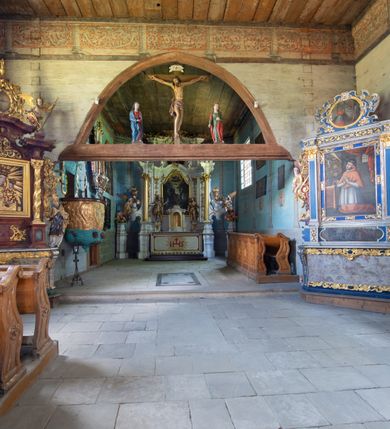 Zdjęcie nr 13: Kościół jest usytuowany ustronnie na stoku wzniesienia, otoczony starodrzewiem i parkanem, ujmującym stary cmentarz z nielicznymi mogiłami i nagrobkami. Orientowaną świątynię w całości wzniesiono z drewna. Zastosowano konstrukcję zrębową na murowanej podwalinie z murowaną kryptą pod prezbiterium. Nawa kościoła jest prawie kwadratowa. Od wchodu przylega do niej węższe prezbiterium zamknięte trójbocznie. Przy jego północnej ścianie wzniesiono bezokienną zakrystię z wejściem od strony wschodniej. Od zachodu z nawą łączy się wieża na rzucie kwadratu wzniesiona na tzw. słup. Ma pochyłe ściany, szalowane w dolnej części deskami, a w górnej podbite gontem. Wieża zakończona jest nadwieszoną izbicą, zwieńczoną ostrosłupowym hełmem gontowym. W połowie wysokości wieżę opasa pulpitowy daszek gontowy łączący się po bokach z dachem nawy. Pod wieżą urządzono obszerną kruchtę. Wysokie i spadziste dachy kościoła pokryte są gontem. Więźba ma konstrukcję storczykową. Ponad wschodnim szczytem nawy usytuowano nadwieszoną wieżyczkę na sygnaturkę o kształcie cebulastym z latarnią zwieńczoną krzyżem. Całość bryły kościoła (z wyjątkiem wieży) obiegają otwarte soboty, w które przed zakrystią od wschodu wbudowano nowy przedsionek. W nawie od południa znajduje się profilowany portal z nadprożem wykrojonym w ośli grzbiet. Umieszczone w nim drzwi zostały zaopatrzone w stare okucia i antabę. Wnętrze nawy i prezbiterium nakryto płaskimi stropami z pozostałościami późnorenesansowych malowideł z motywem kasetonów z rozetami. Łuk tęczowy jest ostry. Spływa na profilowaną belkę, która jest wsparta na kroksztynach. Na belce tęczowej został ustawiony krucyfiks oraz figury Matki Boskiej i św. Jana Apostoła. Chór muzyczny z dekorowanym malarsko parapetem został wsparty na dwóch słupach. W świątyni znajduje się rokokowy ołtarz główny z obrazem ze sceną Zwiastowania NMP. Dwa ołtarze boczne ustawione zostały przy łuku tęczowym. Prawy ołtarz boczny św. Karola Boromeusza pochodzi z drugiej połowy XVII wieku. Na to miejsce trafił on w połowie XX wieku z kaplicy pałacu w Osieku. Lewy ołtarz boczny jest późnobarokowy z około 1700 roku. W jego centrum umieszczono obraz Matki Boskiej Piekarskiej. Na północnej ścianie nad wejściem do zakrystii zawieszono epitafium Mikołaja i Beaty z Mirowa Porębskich z 1611 roku. Wcześniej epitafium pełniło rolę retabulum w miejscu stojącego dziś ołtarza św. Karola Boromeusza. W prezbiterium przy łuku tęczowym zawieszono rokokową ambonę w kształcie łodzi, na którą wejście prowadzi z zakrystii. Przy wejściu do nawy znajduje się kamienna chrzcielnica z początku XVI wieku, ozdobiona herbem Fogelweder, z drewnianą, późnobarokową nakrywą. W zakrystii zachował się także rokokowy drewniany lawetarz z drugiej połowy XVIII wieku.     