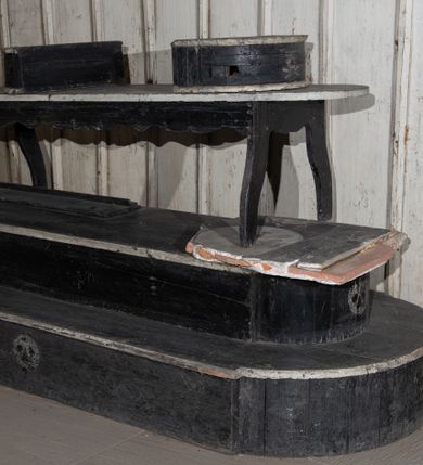 Zdjęcie nr 1: Malowany na czarno katafalk z listwami dekorowanymi białą farbą składa się z kilku części. Jego podstawę tworzą dwa zmniejszające się stopnie na rzucie wydłużonego prostokąta o półkoliście zamkniętych, nadwieszonych krótszych bokach. Dłuższe boki dolnego stopnia i krótsze boki górnego stopnia zdobi malowane na biało wyobrażenie stylizowanej trupiej czaszki. Na górnym stopniu ustawiono ławę malowaną na czarno, wspartą na wyciętych falisto w desce czterech nogach z białym blatem na rzucie wydłużonego prostokąta o półokrągłych krótszych bokach. Na blacie znajdują się dwie półokrągłe czarne podstawy pod trumnę, dekorowane od przodu malowaną na biało stylizowaną trupią czaszką i skrzyżowanymi piszczelami. 