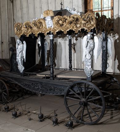 Zdjęcie nr 3: Konny karawan uzyskał formę czterokołowego wozu na resorach z baldachimem wspartym na czterech filarach (w osiach skrajnych) i dwóch kolumnach o skręconych trzonach (w osiach środkowych). Na filarach umieszczono płaskorzeźbione i srebrzone półpostacie lamentujących aniołów, wsparte na wolutowych konsolach dekorowanych palmetami i akantem. Długowłose anioły ukazane zostały w wąskich tunikach przewiązanych w pasie. W lewej ręce trzymają wieńce, prawą ręką ugiętą w łokciu dotykają twarzy w geście rozpaczy. Baldachim przyjął kształt czterospadowego dachu zwieńczonego krzyżem. Spływy dachu ozdobiono postrzępionymi, srebrzonymi liśćmi palmowymi. Jego boki dekoruje płaskorzeźbiona, ażurowa i złocona stylizowana wić akantowa, złączona w dolnej części z arkaturą. Na dłuższych bokach baldachimu ornament rozdziela z jednej strony srebrzona i płaskorzeźbiona głowa Chrystusa w koronie cierniowej w typie Veraikon, z drugiej główki anielskiej oraz zachowana częściowo głowa Matki Boskiej Bolesnej. Dodatkowo płaskorzeźbiony ornament roślinno-kwiatowy zdobi dolne, dłuższe boki karawanu. Z przodu karawanu usytuowano siedzisko w formie płaskorzeźbionego fotela o wolutowo skręconych podłokietnikach. Zaplecek zwieńczony został płaskorzeźbionym ornamentem akantowym, a boki fotela udekorowano płaskorzeźbionymi liśćmi dębu. Odwrocie zaplecek ozdobiono płaskorzeźbionymi liśćmi akantu, wieńcem z liści dębowych oraz dwoma skrzyżowanymi liśćmi palmowymi. Przed fotelem znajduje się klapa na oparcie nóg dla woźnicy. Pod nią usytuowano drewniane elementy pozwalające zaprzęgnąć konia. Drewniane i okute koła z przodu są mniejsze od kół z tyłu.  