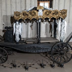 Zdjęcie nr 1: Konny karawan uzyskał formę czterokołowego wozu na resorach z baldachimem wspartym na czterech filarach (w osiach skrajnych) i dwóch kolumnach o skręconych trzonach (w osiach środkowych). Na filarach umieszczono płaskorzeźbione i srebrzone półpostacie lamentujących aniołów, wsparte na wolutowych konsolach dekorowanych palmetami i akantem. Długowłose anioły ukazane zostały w wąskich tunikach przewiązanych w pasie. W lewej ręce trzymają wieńce, prawą ręką ugiętą w łokciu dotykają twarzy w geście rozpaczy. Baldachim przyjął kształt czterospadowego dachu zwieńczonego krzyżem. Spływy dachu ozdobiono postrzępionymi, srebrzonymi liśćmi palmowymi. Jego boki dekoruje płaskorzeźbiona, ażurowa i złocona stylizowana wić akantowa, złączona w dolnej części z arkaturą. Na dłuższych bokach baldachimu ornament rozdziela z jednej strony srebrzona i płaskorzeźbiona głowa Chrystusa w koronie cierniowej w typie Veraikon, z drugiej główki anielskiej oraz zachowana częściowo głowa Matki Boskiej Bolesnej. Dodatkowo płaskorzeźbiony ornament roślinno-kwiatowy zdobi dolne, dłuższe boki karawanu. Z przodu karawanu usytuowano siedzisko w formie płaskorzeźbionego fotela o wolutowo skręconych podłokietnikach. Zaplecek zwieńczony został płaskorzeźbionym ornamentem akantowym, a boki fotela udekorowano płaskorzeźbionymi liśćmi dębu. Odwrocie zaplecek ozdobiono płaskorzeźbionymi liśćmi akantu, wieńcem z liści dębowych oraz dwoma skrzyżowanymi liśćmi palmowymi. Przed fotelem znajduje się klapa na oparcie nóg dla woźnicy. Pod nią usytuowano drewniane elementy pozwalające zaprzęgnąć konia. Drewniane i okute koła z przodu są mniejsze od kół z tyłu.  