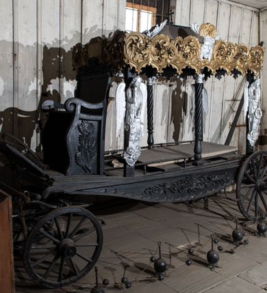 Zdjęcie nr 2: Konny karawan uzyskał formę czterokołowego wozu na resorach z baldachimem wspartym na czterech filarach (w osiach skrajnych) i dwóch kolumnach o skręconych trzonach (w osiach środkowych). Na filarach umieszczono płaskorzeźbione i srebrzone półpostacie lamentujących aniołów, wsparte na wolutowych konsolach dekorowanych palmetami i akantem. Długowłose anioły ukazane zostały w wąskich tunikach przewiązanych w pasie. W lewej ręce trzymają wieńce, prawą ręką ugiętą w łokciu dotykają twarzy w geście rozpaczy. Baldachim przyjął kształt czterospadowego dachu zwieńczonego krzyżem. Spływy dachu ozdobiono postrzępionymi, srebrzonymi liśćmi palmowymi. Jego boki dekoruje płaskorzeźbiona, ażurowa i złocona stylizowana wić akantowa, złączona w dolnej części z arkaturą. Na dłuższych bokach baldachimu ornament rozdziela z jednej strony srebrzona i płaskorzeźbiona głowa Chrystusa w koronie cierniowej w typie Veraikon, z drugiej główki anielskiej oraz zachowana częściowo głowa Matki Boskiej Bolesnej. Dodatkowo płaskorzeźbiony ornament roślinno-kwiatowy zdobi dolne, dłuższe boki karawanu. Z przodu karawanu usytuowano siedzisko w formie płaskorzeźbionego fotela o wolutowo skręconych podłokietnikach. Zaplecek zwieńczony został płaskorzeźbionym ornamentem akantowym, a boki fotela udekorowano płaskorzeźbionymi liśćmi dębu. Odwrocie zaplecek ozdobiono płaskorzeźbionymi liśćmi akantu, wieńcem z liści dębowych oraz dwoma skrzyżowanymi liśćmi palmowymi. Przed fotelem znajduje się klapa na oparcie nóg dla woźnicy. Pod nią usytuowano drewniane elementy pozwalające zaprzęgnąć konia. Drewniane i okute koła z przodu są mniejsze od kół z tyłu.  