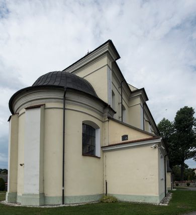 Zdjęcie nr 17: Kościół jest położony w centrum wsi, na wschód od głównej drogi, na placu otoczonym kamiennym murem przykrytym dachówką, z kilkoma drzewami. Wejście na plac przykościelny prowadzi przez szerokie schody i bramę od zachodu i bramkę od północy. Kościół jest budowlą założoną na planie prostokąta, jednonawowy, trójprzęsłowy z węższym, dwuprzęsłowym prezbiterium z apsydą. Do nawy przylegają od zachodu i północy dwie kruchty na planie kwadratu i wieża w narożu południowo-zachodnim fasady. Do prezbiterium od północy jest dostawiona kaplica na rzucie prostokąta otwierająca się półkolistymi arkadami, a od południa zakrystia. 
Artykulacja ścian jest przeprowadzona za pomocą zdwojonych pilastrów toskańskich stojących na wysokich postumentach i podtrzymujących wydatne, przełamujące się, pełne belkowanie z fryzem tryglifowym oraz wysuniętym gzymsem, obiegającym całe wnętrze świątyni. Wnętrze nakrywa sklepienie kolebkowe na gurtach z lunetami; w kluczach sklepienia znajdują się tarcze herbowe, Złota Pilawa oraz Ostoja. W lunetach umieszczone są okna prostokątne zamknięte łukiem odcinkowym; takie same okna znajdują się w apsydzie prezbiterium, poniżej belkowania.  W kaplicy znajdują się dwa prostokątne okna zamknięte odcinkowo. Zakrystia jest jednoprzestrzenna, nakryta sklepieniem krzyżowym. Prowadzi do niej kamienny portal profilowany z uszakami, z podwieszonymi wolutami, zwieńczony wydatnym gzymsem. W partii fryzu umieszczono polichromowany kartusz z herbem Ostoja. W ścianach środkowego przęsła korpusu nawowego po obu stronach kościoła znajdują się głębokie, prostokątne nisze zamknięte łukami odcinkowymi. W niszy północnej umieszczono drzwi prowadzące do niewielkiej kruchty przykrytej stropem, z dwoma małymi, okrągłymi oknami po bokach. Również po zachodniej stronie korpusu znajduje się wejście do kruchty. Ponadto na ścianach nawy oraz prezbiterium widnieją płyciny ujęte ramą profilowaną w tynku. W pierwszym od strony fasady przęśle znajduje się murowany chór muzyczny, trójarkadowy, wsparty na dwóch filarach opiętych pilastrami oraz dwóch półfilarach przylegających do ścian, sklepiony krzyżowo. 
Fasada kościoła jest płaska, zakończona gzymsem, z czterokondygnacyjną wieżą, nakrytą cebulastym hełmem z krzyżem. Boczne elewacje ujęte uskokowymi przyporami, zakończone gzymsem. Dachy dwuspadowe nad nawą i kruchtami, trójspadowe nad prezbiterium, półsferyczne nad apsydą, pulpitowe nad kaplicą i zakrystią, kryte blachą. Nad granicy nawy i prezbiterium wieloboczna wieżyczka na sygnaturkę z prześwitami, zakończona kopułą, sterczyną i kulą z krzyżem. 
