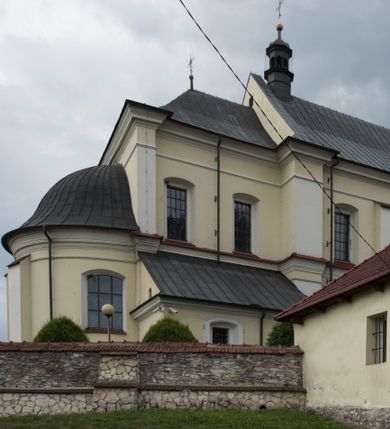 Zdjęcie nr 14: Kościół jest położony w centrum wsi, na wschód od głównej drogi, na placu otoczonym kamiennym murem przykrytym dachówką, z kilkoma drzewami. Wejście na plac przykościelny prowadzi przez szerokie schody i bramę od zachodu i bramkę od północy. Kościół jest budowlą założoną na planie prostokąta, jednonawowy, trójprzęsłowy z węższym, dwuprzęsłowym prezbiterium z apsydą. Do nawy przylegają od zachodu i północy dwie kruchty na planie kwadratu i wieża w narożu południowo-zachodnim fasady. Do prezbiterium od północy jest dostawiona kaplica na rzucie prostokąta otwierająca się półkolistymi arkadami, a od południa zakrystia. 
Artykulacja ścian jest przeprowadzona za pomocą zdwojonych pilastrów toskańskich stojących na wysokich postumentach i podtrzymujących wydatne, przełamujące się, pełne belkowanie z fryzem tryglifowym oraz wysuniętym gzymsem, obiegającym całe wnętrze świątyni. Wnętrze nakrywa sklepienie kolebkowe na gurtach z lunetami; w kluczach sklepienia znajdują się tarcze herbowe, Złota Pilawa oraz Ostoja. W lunetach umieszczone są okna prostokątne zamknięte łukiem odcinkowym; takie same okna znajdują się w apsydzie prezbiterium, poniżej belkowania.  W kaplicy znajdują się dwa prostokątne okna zamknięte odcinkowo. Zakrystia jest jednoprzestrzenna, nakryta sklepieniem krzyżowym. Prowadzi do niej kamienny portal profilowany z uszakami, z podwieszonymi wolutami, zwieńczony wydatnym gzymsem. W partii fryzu umieszczono polichromowany kartusz z herbem Ostoja. W ścianach środkowego przęsła korpusu nawowego po obu stronach kościoła znajdują się głębokie, prostokątne nisze zamknięte łukami odcinkowymi. W niszy północnej umieszczono drzwi prowadzące do niewielkiej kruchty przykrytej stropem, z dwoma małymi, okrągłymi oknami po bokach. Również po zachodniej stronie korpusu znajduje się wejście do kruchty. Ponadto na ścianach nawy oraz prezbiterium widnieją płyciny ujęte ramą profilowaną w tynku. W pierwszym od strony fasady przęśle znajduje się murowany chór muzyczny, trójarkadowy, wsparty na dwóch filarach opiętych pilastrami oraz dwóch półfilarach przylegających do ścian, sklepiony krzyżowo. 
Fasada kościoła jest płaska, zakończona gzymsem, z czterokondygnacyjną wieżą, nakrytą cebulastym hełmem z krzyżem. Boczne elewacje ujęte uskokowymi przyporami, zakończone gzymsem. Dachy dwuspadowe nad nawą i kruchtami, trójspadowe nad prezbiterium, półsferyczne nad apsydą, pulpitowe nad kaplicą i zakrystią, kryte blachą. Nad granicy nawy i prezbiterium wieloboczna wieżyczka na sygnaturkę z prześwitami, zakończona kopułą, sterczyną i kulą z krzyżem. 
