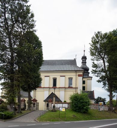 Zdjęcie nr 25: Kościół jest położony w centrum wsi, na wschód od głównej drogi, na placu otoczonym kamiennym murem przykrytym dachówką, z kilkoma drzewami. Wejście na plac przykościelny prowadzi przez szerokie schody i bramę od zachodu i bramkę od północy. Kościół jest budowlą założoną na planie prostokąta, jednonawowy, trójprzęsłowy z węższym, dwuprzęsłowym prezbiterium z apsydą. Do nawy przylegają od zachodu i północy dwie kruchty na planie kwadratu i wieża w narożu południowo-zachodnim fasady. Do prezbiterium od północy jest dostawiona kaplica na rzucie prostokąta otwierająca się półkolistymi arkadami, a od południa zakrystia. 
Artykulacja ścian jest przeprowadzona za pomocą zdwojonych pilastrów toskańskich stojących na wysokich postumentach i podtrzymujących wydatne, przełamujące się, pełne belkowanie z fryzem tryglifowym oraz wysuniętym gzymsem, obiegającym całe wnętrze świątyni. Wnętrze nakrywa sklepienie kolebkowe na gurtach z lunetami; w kluczach sklepienia znajdują się tarcze herbowe, Złota Pilawa oraz Ostoja. W lunetach umieszczone są okna prostokątne zamknięte łukiem odcinkowym; takie same okna znajdują się w apsydzie prezbiterium, poniżej belkowania.  W kaplicy znajdują się dwa prostokątne okna zamknięte odcinkowo. Zakrystia jest jednoprzestrzenna, nakryta sklepieniem krzyżowym. Prowadzi do niej kamienny portal profilowany z uszakami, z podwieszonymi wolutami, zwieńczony wydatnym gzymsem. W partii fryzu umieszczono polichromowany kartusz z herbem Ostoja. W ścianach środkowego przęsła korpusu nawowego po obu stronach kościoła znajdują się głębokie, prostokątne nisze zamknięte łukami odcinkowymi. W niszy północnej umieszczono drzwi prowadzące do niewielkiej kruchty przykrytej stropem, z dwoma małymi, okrągłymi oknami po bokach. Również po zachodniej stronie korpusu znajduje się wejście do kruchty. Ponadto na ścianach nawy oraz prezbiterium widnieją płyciny ujęte ramą profilowaną w tynku. W pierwszym od strony fasady przęśle znajduje się murowany chór muzyczny, trójarkadowy, wsparty na dwóch filarach opiętych pilastrami oraz dwóch półfilarach przylegających do ścian, sklepiony krzyżowo. 
Fasada kościoła jest płaska, zakończona gzymsem, z czterokondygnacyjną wieżą, nakrytą cebulastym hełmem z krzyżem. Boczne elewacje ujęte uskokowymi przyporami, zakończone gzymsem. Dachy dwuspadowe nad nawą i kruchtami, trójspadowe nad prezbiterium, półsferyczne nad apsydą, pulpitowe nad kaplicą i zakrystią, kryte blachą. Nad granicy nawy i prezbiterium wieloboczna wieżyczka na sygnaturkę z prześwitami, zakończona kopułą, sterczyną i kulą z krzyżem. 
