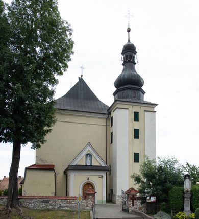 Zdjęcie nr 27: Kościół jest położony w centrum wsi, na wschód od głównej drogi, na placu otoczonym kamiennym murem przykrytym dachówką, z kilkoma drzewami. Wejście na plac przykościelny prowadzi przez szerokie schody i bramę od zachodu i bramkę od północy. Kościół jest budowlą założoną na planie prostokąta, jednonawowy, trójprzęsłowy z węższym, dwuprzęsłowym prezbiterium z apsydą. Do nawy przylegają od zachodu i północy dwie kruchty na planie kwadratu i wieża w narożu południowo-zachodnim fasady. Do prezbiterium od północy jest dostawiona kaplica na rzucie prostokąta otwierająca się półkolistymi arkadami, a od południa zakrystia. 
Artykulacja ścian jest przeprowadzona za pomocą zdwojonych pilastrów toskańskich stojących na wysokich postumentach i podtrzymujących wydatne, przełamujące się, pełne belkowanie z fryzem tryglifowym oraz wysuniętym gzymsem, obiegającym całe wnętrze świątyni. Wnętrze nakrywa sklepienie kolebkowe na gurtach z lunetami; w kluczach sklepienia znajdują się tarcze herbowe, Złota Pilawa oraz Ostoja. W lunetach umieszczone są okna prostokątne zamknięte łukiem odcinkowym; takie same okna znajdują się w apsydzie prezbiterium, poniżej belkowania.  W kaplicy znajdują się dwa prostokątne okna zamknięte odcinkowo. Zakrystia jest jednoprzestrzenna, nakryta sklepieniem krzyżowym. Prowadzi do niej kamienny portal profilowany z uszakami, z podwieszonymi wolutami, zwieńczony wydatnym gzymsem. W partii fryzu umieszczono polichromowany kartusz z herbem Ostoja. W ścianach środkowego przęsła korpusu nawowego po obu stronach kościoła znajdują się głębokie, prostokątne nisze zamknięte łukami odcinkowymi. W niszy północnej umieszczono drzwi prowadzące do niewielkiej kruchty przykrytej stropem, z dwoma małymi, okrągłymi oknami po bokach. Również po zachodniej stronie korpusu znajduje się wejście do kruchty. Ponadto na ścianach nawy oraz prezbiterium widnieją płyciny ujęte ramą profilowaną w tynku. W pierwszym od strony fasady przęśle znajduje się murowany chór muzyczny, trójarkadowy, wsparty na dwóch filarach opiętych pilastrami oraz dwóch półfilarach przylegających do ścian, sklepiony krzyżowo. 
Fasada kościoła jest płaska, zakończona gzymsem, z czterokondygnacyjną wieżą, nakrytą cebulastym hełmem z krzyżem. Boczne elewacje ujęte uskokowymi przyporami, zakończone gzymsem. Dachy dwuspadowe nad nawą i kruchtami, trójspadowe nad prezbiterium, półsferyczne nad apsydą, pulpitowe nad kaplicą i zakrystią, kryte blachą. Nad granicy nawy i prezbiterium wieloboczna wieżyczka na sygnaturkę z prześwitami, zakończona kopułą, sterczyną i kulą z krzyżem. 
