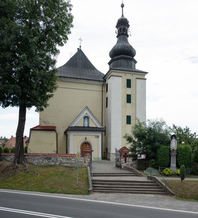 Zdjęcie nr 4: Kościół jest położony w centrum wsi, na wschód od głównej drogi, na placu otoczonym kamiennym murem przykrytym dachówką, z kilkoma drzewami. Wejście na plac przykościelny prowadzi przez szerokie schody i bramę od zachodu i bramkę od północy. Kościół jest budowlą założoną na planie prostokąta, jednonawowy, trójprzęsłowy z węższym, dwuprzęsłowym prezbiterium z apsydą. Do nawy przylegają od zachodu i północy dwie kruchty na planie kwadratu i wieża w narożu południowo-zachodnim fasady. Do prezbiterium od północy jest dostawiona kaplica na rzucie prostokąta otwierająca się półkolistymi arkadami, a od południa zakrystia. 
Artykulacja ścian jest przeprowadzona za pomocą zdwojonych pilastrów toskańskich stojących na wysokich postumentach i podtrzymujących wydatne, przełamujące się, pełne belkowanie z fryzem tryglifowym oraz wysuniętym gzymsem, obiegającym całe wnętrze świątyni. Wnętrze nakrywa sklepienie kolebkowe na gurtach z lunetami; w kluczach sklepienia znajdują się tarcze herbowe, Złota Pilawa oraz Ostoja. W lunetach umieszczone są okna prostokątne zamknięte łukiem odcinkowym; takie same okna znajdują się w apsydzie prezbiterium, poniżej belkowania.  W kaplicy znajdują się dwa prostokątne okna zamknięte odcinkowo. Zakrystia jest jednoprzestrzenna, nakryta sklepieniem krzyżowym. Prowadzi do niej kamienny portal profilowany z uszakami, z podwieszonymi wolutami, zwieńczony wydatnym gzymsem. W partii fryzu umieszczono polichromowany kartusz z herbem Ostoja. W ścianach środkowego przęsła korpusu nawowego po obu stronach kościoła znajdują się głębokie, prostokątne nisze zamknięte łukami odcinkowymi. W niszy północnej umieszczono drzwi prowadzące do niewielkiej kruchty przykrytej stropem, z dwoma małymi, okrągłymi oknami po bokach. Również po zachodniej stronie korpusu znajduje się wejście do kruchty. Ponadto na ścianach nawy oraz prezbiterium widnieją płyciny ujęte ramą profilowaną w tynku. W pierwszym od strony fasady przęśle znajduje się murowany chór muzyczny, trójarkadowy, wsparty na dwóch filarach opiętych pilastrami oraz dwóch półfilarach przylegających do ścian, sklepiony krzyżowo. 
Fasada kościoła jest płaska, zakończona gzymsem, z czterokondygnacyjną wieżą, nakrytą cebulastym hełmem z krzyżem. Boczne elewacje ujęte uskokowymi przyporami, zakończone gzymsem. Dachy dwuspadowe nad nawą i kruchtami, trójspadowe nad prezbiterium, półsferyczne nad apsydą, pulpitowe nad kaplicą i zakrystią, kryte blachą. Nad granicy nawy i prezbiterium wieloboczna wieżyczka na sygnaturkę z prześwitami, zakończona kopułą, sterczyną i kulą z krzyżem. 
