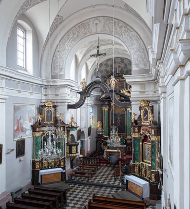 Zdjęcie nr 7: Kościół jest położony w centrum wsi, na wschód od głównej drogi, na placu otoczonym kamiennym murem przykrytym dachówką, z kilkoma drzewami. Wejście na plac przykościelny prowadzi przez szerokie schody i bramę od zachodu i bramkę od północy. Kościół jest budowlą założoną na planie prostokąta, jednonawowy, trójprzęsłowy z węższym, dwuprzęsłowym prezbiterium z apsydą. Do nawy przylegają od zachodu i północy dwie kruchty na planie kwadratu i wieża w narożu południowo-zachodnim fasady. Do prezbiterium od północy jest dostawiona kaplica na rzucie prostokąta otwierająca się półkolistymi arkadami, a od południa zakrystia. 
Artykulacja ścian jest przeprowadzona za pomocą zdwojonych pilastrów toskańskich stojących na wysokich postumentach i podtrzymujących wydatne, przełamujące się, pełne belkowanie z fryzem tryglifowym oraz wysuniętym gzymsem, obiegającym całe wnętrze świątyni. Wnętrze nakrywa sklepienie kolebkowe na gurtach z lunetami; w kluczach sklepienia znajdują się tarcze herbowe, Złota Pilawa oraz Ostoja. W lunetach umieszczone są okna prostokątne zamknięte łukiem odcinkowym; takie same okna znajdują się w apsydzie prezbiterium, poniżej belkowania.  W kaplicy znajdują się dwa prostokątne okna zamknięte odcinkowo. Zakrystia jest jednoprzestrzenna, nakryta sklepieniem krzyżowym. Prowadzi do niej kamienny portal profilowany z uszakami, z podwieszonymi wolutami, zwieńczony wydatnym gzymsem. W partii fryzu umieszczono polichromowany kartusz z herbem Ostoja. W ścianach środkowego przęsła korpusu nawowego po obu stronach kościoła znajdują się głębokie, prostokątne nisze zamknięte łukami odcinkowymi. W niszy północnej umieszczono drzwi prowadzące do niewielkiej kruchty przykrytej stropem, z dwoma małymi, okrągłymi oknami po bokach. Również po zachodniej stronie korpusu znajduje się wejście do kruchty. Ponadto na ścianach nawy oraz prezbiterium widnieją płyciny ujęte ramą profilowaną w tynku. W pierwszym od strony fasady przęśle znajduje się murowany chór muzyczny, trójarkadowy, wsparty na dwóch filarach opiętych pilastrami oraz dwóch półfilarach przylegających do ścian, sklepiony krzyżowo. 
Fasada kościoła jest płaska, zakończona gzymsem, z czterokondygnacyjną wieżą, nakrytą cebulastym hełmem z krzyżem. Boczne elewacje ujęte uskokowymi przyporami, zakończone gzymsem. Dachy dwuspadowe nad nawą i kruchtami, trójspadowe nad prezbiterium, półsferyczne nad apsydą, pulpitowe nad kaplicą i zakrystią, kryte blachą. Nad granicy nawy i prezbiterium wieloboczna wieżyczka na sygnaturkę z prześwitami, zakończona kopułą, sterczyną i kulą z krzyżem. 
