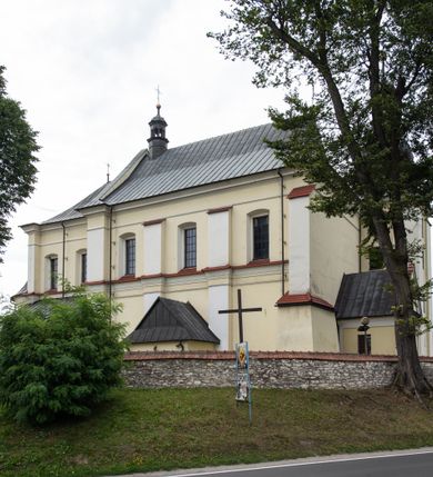 Zdjęcie nr 6: Kościół jest położony w centrum wsi, na wschód od głównej drogi, na placu otoczonym kamiennym murem przykrytym dachówką, z kilkoma drzewami. Wejście na plac przykościelny prowadzi przez szerokie schody i bramę od zachodu i bramkę od północy. Kościół jest budowlą założoną na planie prostokąta, jednonawowy, trójprzęsłowy z węższym, dwuprzęsłowym prezbiterium z apsydą. Do nawy przylegają od zachodu i północy dwie kruchty na planie kwadratu i wieża w narożu południowo-zachodnim fasady. Do prezbiterium od północy jest dostawiona kaplica na rzucie prostokąta otwierająca się półkolistymi arkadami, a od południa zakrystia. 
Artykulacja ścian jest przeprowadzona za pomocą zdwojonych pilastrów toskańskich stojących na wysokich postumentach i podtrzymujących wydatne, przełamujące się, pełne belkowanie z fryzem tryglifowym oraz wysuniętym gzymsem, obiegającym całe wnętrze świątyni. Wnętrze nakrywa sklepienie kolebkowe na gurtach z lunetami; w kluczach sklepienia znajdują się tarcze herbowe, Złota Pilawa oraz Ostoja. W lunetach umieszczone są okna prostokątne zamknięte łukiem odcinkowym; takie same okna znajdują się w apsydzie prezbiterium, poniżej belkowania.  W kaplicy znajdują się dwa prostokątne okna zamknięte odcinkowo. Zakrystia jest jednoprzestrzenna, nakryta sklepieniem krzyżowym. Prowadzi do niej kamienny portal profilowany z uszakami, z podwieszonymi wolutami, zwieńczony wydatnym gzymsem. W partii fryzu umieszczono polichromowany kartusz z herbem Ostoja. W ścianach środkowego przęsła korpusu nawowego po obu stronach kościoła znajdują się głębokie, prostokątne nisze zamknięte łukami odcinkowymi. W niszy północnej umieszczono drzwi prowadzące do niewielkiej kruchty przykrytej stropem, z dwoma małymi, okrągłymi oknami po bokach. Również po zachodniej stronie korpusu znajduje się wejście do kruchty. Ponadto na ścianach nawy oraz prezbiterium widnieją płyciny ujęte ramą profilowaną w tynku. W pierwszym od strony fasady przęśle znajduje się murowany chór muzyczny, trójarkadowy, wsparty na dwóch filarach opiętych pilastrami oraz dwóch półfilarach przylegających do ścian, sklepiony krzyżowo. 
Fasada kościoła jest płaska, zakończona gzymsem, z czterokondygnacyjną wieżą, nakrytą cebulastym hełmem z krzyżem. Boczne elewacje ujęte uskokowymi przyporami, zakończone gzymsem. Dachy dwuspadowe nad nawą i kruchtami, trójspadowe nad prezbiterium, półsferyczne nad apsydą, pulpitowe nad kaplicą i zakrystią, kryte blachą. Nad granicy nawy i prezbiterium wieloboczna wieżyczka na sygnaturkę z prześwitami, zakończona kopułą, sterczyną i kulą z krzyżem. 
