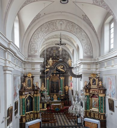 Zdjęcie nr 5: Kościół jest położony w centrum wsi, na wschód od głównej drogi, na placu otoczonym kamiennym murem przykrytym dachówką, z kilkoma drzewami. Wejście na plac przykościelny prowadzi przez szerokie schody i bramę od zachodu i bramkę od północy. Kościół jest budowlą założoną na planie prostokąta, jednonawowy, trójprzęsłowy z węższym, dwuprzęsłowym prezbiterium z apsydą. Do nawy przylegają od zachodu i północy dwie kruchty na planie kwadratu i wieża w narożu południowo-zachodnim fasady. Do prezbiterium od północy jest dostawiona kaplica na rzucie prostokąta otwierająca się półkolistymi arkadami, a od południa zakrystia. 
Artykulacja ścian jest przeprowadzona za pomocą zdwojonych pilastrów toskańskich stojących na wysokich postumentach i podtrzymujących wydatne, przełamujące się, pełne belkowanie z fryzem tryglifowym oraz wysuniętym gzymsem, obiegającym całe wnętrze świątyni. Wnętrze nakrywa sklepienie kolebkowe na gurtach z lunetami; w kluczach sklepienia znajdują się tarcze herbowe, Złota Pilawa oraz Ostoja. W lunetach umieszczone są okna prostokątne zamknięte łukiem odcinkowym; takie same okna znajdują się w apsydzie prezbiterium, poniżej belkowania.  W kaplicy znajdują się dwa prostokątne okna zamknięte odcinkowo. Zakrystia jest jednoprzestrzenna, nakryta sklepieniem krzyżowym. Prowadzi do niej kamienny portal profilowany z uszakami, z podwieszonymi wolutami, zwieńczony wydatnym gzymsem. W partii fryzu umieszczono polichromowany kartusz z herbem Ostoja. W ścianach środkowego przęsła korpusu nawowego po obu stronach kościoła znajdują się głębokie, prostokątne nisze zamknięte łukami odcinkowymi. W niszy północnej umieszczono drzwi prowadzące do niewielkiej kruchty przykrytej stropem, z dwoma małymi, okrągłymi oknami po bokach. Również po zachodniej stronie korpusu znajduje się wejście do kruchty. Ponadto na ścianach nawy oraz prezbiterium widnieją płyciny ujęte ramą profilowaną w tynku. W pierwszym od strony fasady przęśle znajduje się murowany chór muzyczny, trójarkadowy, wsparty na dwóch filarach opiętych pilastrami oraz dwóch półfilarach przylegających do ścian, sklepiony krzyżowo. 
Fasada kościoła jest płaska, zakończona gzymsem, z czterokondygnacyjną wieżą, nakrytą cebulastym hełmem z krzyżem. Boczne elewacje ujęte uskokowymi przyporami, zakończone gzymsem. Dachy dwuspadowe nad nawą i kruchtami, trójspadowe nad prezbiterium, półsferyczne nad apsydą, pulpitowe nad kaplicą i zakrystią, kryte blachą. Nad granicy nawy i prezbiterium wieloboczna wieżyczka na sygnaturkę z prześwitami, zakończona kopułą, sterczyną i kulą z krzyżem. 
