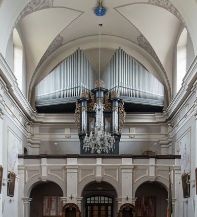 Zdjęcie nr 12: Kościół jest położony w centrum wsi, na wschód od głównej drogi, na placu otoczonym kamiennym murem przykrytym dachówką, z kilkoma drzewami. Wejście na plac przykościelny prowadzi przez szerokie schody i bramę od zachodu i bramkę od północy. Kościół jest budowlą założoną na planie prostokąta, jednonawowy, trójprzęsłowy z węższym, dwuprzęsłowym prezbiterium z apsydą. Do nawy przylegają od zachodu i północy dwie kruchty na planie kwadratu i wieża w narożu południowo-zachodnim fasady. Do prezbiterium od północy jest dostawiona kaplica na rzucie prostokąta otwierająca się półkolistymi arkadami, a od południa zakrystia. 
Artykulacja ścian jest przeprowadzona za pomocą zdwojonych pilastrów toskańskich stojących na wysokich postumentach i podtrzymujących wydatne, przełamujące się, pełne belkowanie z fryzem tryglifowym oraz wysuniętym gzymsem, obiegającym całe wnętrze świątyni. Wnętrze nakrywa sklepienie kolebkowe na gurtach z lunetami; w kluczach sklepienia znajdują się tarcze herbowe, Złota Pilawa oraz Ostoja. W lunetach umieszczone są okna prostokątne zamknięte łukiem odcinkowym; takie same okna znajdują się w apsydzie prezbiterium, poniżej belkowania.  W kaplicy znajdują się dwa prostokątne okna zamknięte odcinkowo. Zakrystia jest jednoprzestrzenna, nakryta sklepieniem krzyżowym. Prowadzi do niej kamienny portal profilowany z uszakami, z podwieszonymi wolutami, zwieńczony wydatnym gzymsem. W partii fryzu umieszczono polichromowany kartusz z herbem Ostoja. W ścianach środkowego przęsła korpusu nawowego po obu stronach kościoła znajdują się głębokie, prostokątne nisze zamknięte łukami odcinkowymi. W niszy północnej umieszczono drzwi prowadzące do niewielkiej kruchty przykrytej stropem, z dwoma małymi, okrągłymi oknami po bokach. Również po zachodniej stronie korpusu znajduje się wejście do kruchty. Ponadto na ścianach nawy oraz prezbiterium widnieją płyciny ujęte ramą profilowaną w tynku. W pierwszym od strony fasady przęśle znajduje się murowany chór muzyczny, trójarkadowy, wsparty na dwóch filarach opiętych pilastrami oraz dwóch półfilarach przylegających do ścian, sklepiony krzyżowo. 
Fasada kościoła jest płaska, zakończona gzymsem, z czterokondygnacyjną wieżą, nakrytą cebulastym hełmem z krzyżem. Boczne elewacje ujęte uskokowymi przyporami, zakończone gzymsem. Dachy dwuspadowe nad nawą i kruchtami, trójspadowe nad prezbiterium, półsferyczne nad apsydą, pulpitowe nad kaplicą i zakrystią, kryte blachą. Nad granicy nawy i prezbiterium wieloboczna wieżyczka na sygnaturkę z prześwitami, zakończona kopułą, sterczyną i kulą z krzyżem. 
