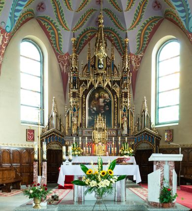 Zdjęcie nr 3: Obraz w kształcie prostokąta stojącego z przedstawieniem Matki Boskiej z Dzieciątkiem w typie Eleusy. W centrum ukazana Matka Boska w półpostaci, zwrócona delikatnie w lewo, z pochyloną głową w stronę Dzieciątka, które trzyma na rękach. Twarz owalna o mocno zaznaczonych rysach nosa i łuków brwiowych, wzrok skierowany na wprost. Ubrana w czerwoną suknię ze złotą lamówką, obficie drapowaną oraz granatowy maforion ze złotą lamówką zarzucony na głowę i ramiona z gwiazdą na prawym ramieniu. Dzieciątko Jezus ukazane z lewego profilu, przytula twarz do twarzy Matki Boskiej, prawą ręką ujmuje jej brodę, a lewą szyję. Ma twarz owalną o wyrazistych rysach. Ubrane jest w białą szatę z rękawami podwiniętymi do łokci. Wokół głowy Matki Boskiej duży złoto-czerwony nimb. Tło jednolite, granatowe. Kolorystyka intensywna, silny linearyzm.
