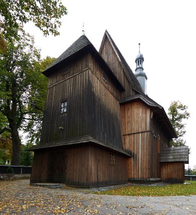 Zdjęcie nr 3: Kościół drewniany, orientowany, usytuowany na wzgórku, otoczony pierwotnym cmentarzem przykościelnym oraz starodrzewiem (lipami), ujęty drewnianym ogrodzeniem z poziomo ułożonych desek, w którego ciągu znajduje się masywna dzwonnica. Ściany kościoła wzniesione w konstrukcji zrębowej, oszalowanej pionowo z listwowaniem. Nawa na planie prostokąta; prezbiterium węższe, zamknięte wielobocznie, zbudowane na zrąb; od północy zakrystia na planie prostokąta; od południa późniejsza, dobudowana kruchta. Od zachodu nawę poprzedza niższa wieża, zbudowana w konstrukcji słupowo-ramowej, o pochyłych ścianach. Wnętrze nakryte stropami, w nawie z zaskrzynieniami. Nawa od prezbiterium oddzielona tęczą o łuku półkolistym wspartym na kroksztynach; na belce umieszczone rzeźby z początku XVI wieku przedstawiające grupę Ukrzyżowania: krucyfiks, Matka Boska, św. Jan Ewangelista oraz św. Maria Magdalena. Chór muzyczny wsparty na dwóch kolumnach, barokowy. Wnętrze pokryte polichromią z pierwszej połowy XVII wieku. Pośrodku kościoła oraz bliżej chóru muzycznego znajdują się wejścia do krypt pokryte płytami. Główny portal kościoła umieszczony jest od strony zachodniej, w przedsionku pod wieżą, ukształtowany w formie profilowanej, lekko zaostrzonej arkady przechodzącej w tzw. ośli grzbiet. Portal południowy, w kruchcie zakończony ostrołukiem, z motywem sznura w profilowaniach z pierwotnymi jednoskrzydłowymi drzwiami o gotyckich okuciach. Portal prowadzący z prezbiterium do zakrystii ma bogate profilowanie (zewnętrzny profil z motywem „ząbków”), jest zakończony w tzw. ośli grzbiet z drzwiami żelaznymi, dekorowanymi ukośną kratą. Okna znajdują się od strony południowej, pod dwa w nawie oraz w prezbiterium. Nadokienniki mają wykrój w ośli grzbiet. Ponadto jedno okno o analogicznym układzie w prezbiterium na ścianie wschodniej, częściowo zabudowane, zasłonięte ołtarzem. Dach gontowy o jednej kalenicy, wsparty na kroksztynach z szerokim okapem, o dużym kącie spadku uzyskanym poprzez zastosowaną gotycką więźbę storczykową, pośrodku z wieżyczką na sygnaturkę o barkowym kształcie. Wieża główna nakryta dachem gontowym, trójspadowym.

