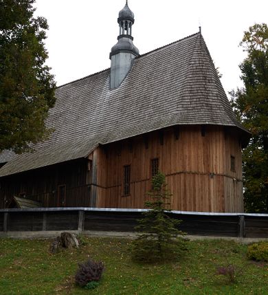 Zdjęcie nr 1: Kościół drewniany, orientowany, usytuowany na wzgórku, otoczony pierwotnym cmentarzem przykościelnym oraz starodrzewiem (lipami), ujęty drewnianym ogrodzeniem z poziomo ułożonych desek, w którego ciągu znajduje się masywna dzwonnica. Ściany kościoła wzniesione w konstrukcji zrębowej, oszalowanej pionowo z listwowaniem. Nawa na planie prostokąta; prezbiterium węższe, zamknięte wielobocznie, zbudowane na zrąb; od północy zakrystia na planie prostokąta; od południa późniejsza, dobudowana kruchta. Od zachodu nawę poprzedza niższa wieża, zbudowana w konstrukcji słupowo-ramowej, o pochyłych ścianach. Wnętrze nakryte stropami, w nawie z zaskrzynieniami. Nawa od prezbiterium oddzielona tęczą o łuku półkolistym wspartym na kroksztynach; na belce umieszczone rzeźby z początku XVI wieku przedstawiające grupę Ukrzyżowania: krucyfiks, Matka Boska, św. Jan Ewangelista oraz św. Maria Magdalena. Chór muzyczny wsparty na dwóch kolumnach, barokowy. Wnętrze pokryte polichromią z pierwszej połowy XVII wieku. Pośrodku kościoła oraz bliżej chóru muzycznego znajdują się wejścia do krypt pokryte płytami. Główny portal kościoła umieszczony jest od strony zachodniej, w przedsionku pod wieżą, ukształtowany w formie profilowanej, lekko zaostrzonej arkady przechodzącej w tzw. ośli grzbiet. Portal południowy, w kruchcie zakończony ostrołukiem, z motywem sznura w profilowaniach z pierwotnymi jednoskrzydłowymi drzwiami o gotyckich okuciach. Portal prowadzący z prezbiterium do zakrystii ma bogate profilowanie (zewnętrzny profil z motywem „ząbków”), jest zakończony w tzw. ośli grzbiet z drzwiami żelaznymi, dekorowanymi ukośną kratą. Okna znajdują się od strony południowej, pod dwa w nawie oraz w prezbiterium. Nadokienniki mają wykrój w ośli grzbiet. Ponadto jedno okno o analogicznym układzie w prezbiterium na ścianie wschodniej, częściowo zabudowane, zasłonięte ołtarzem. Dach gontowy o jednej kalenicy, wsparty na kroksztynach z szerokim okapem, o dużym kącie spadku uzyskanym poprzez zastosowaną gotycką więźbę storczykową, pośrodku z wieżyczką na sygnaturkę o barkowym kształcie. Wieża główna nakryta dachem gontowym, trójspadowym.
