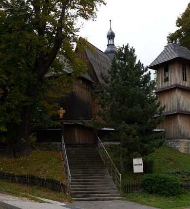 Zdjęcie nr 4: Kościół drewniany, orientowany, usytuowany na wzgórku, otoczony pierwotnym cmentarzem przykościelnym oraz starodrzewiem (lipami), ujęty drewnianym ogrodzeniem z poziomo ułożonych desek, w którego ciągu znajduje się masywna dzwonnica. Ściany kościoła wzniesione w konstrukcji zrębowej, oszalowanej pionowo z listwowaniem. Nawa na planie prostokąta; prezbiterium węższe, zamknięte wielobocznie, zbudowane na zrąb; od północy zakrystia na planie prostokąta; od południa późniejsza, dobudowana kruchta. Od zachodu nawę poprzedza niższa wieża, zbudowana w konstrukcji słupowo-ramowej, o pochyłych ścianach. Wnętrze nakryte stropami, w nawie z zaskrzynieniami. Nawa od prezbiterium oddzielona tęczą o łuku półkolistym wspartym na kroksztynach; na belce umieszczone rzeźby z początku XVI wieku przedstawiające grupę Ukrzyżowania: krucyfiks, Matka Boska, św. Jan Ewangelista oraz św. Maria Magdalena. Chór muzyczny wsparty na dwóch kolumnach, barokowy. Wnętrze pokryte polichromią z pierwszej połowy XVII wieku. Pośrodku kościoła oraz bliżej chóru muzycznego znajdują się wejścia do krypt pokryte płytami. Główny portal kościoła umieszczony jest od strony zachodniej, w przedsionku pod wieżą, ukształtowany w formie profilowanej, lekko zaostrzonej arkady przechodzącej w tzw. ośli grzbiet. Portal południowy, w kruchcie zakończony ostrołukiem, z motywem sznura w profilowaniach z pierwotnymi jednoskrzydłowymi drzwiami o gotyckich okuciach. Portal prowadzący z prezbiterium do zakrystii ma bogate profilowanie (zewnętrzny profil z motywem „ząbków”), jest zakończony w tzw. ośli grzbiet z drzwiami żelaznymi, dekorowanymi ukośną kratą. Okna znajdują się od strony południowej, pod dwa w nawie oraz w prezbiterium. Nadokienniki mają wykrój w ośli grzbiet. Ponadto jedno okno o analogicznym układzie w prezbiterium na ścianie wschodniej, częściowo zabudowane, zasłonięte ołtarzem. Dach gontowy o jednej kalenicy, wsparty na kroksztynach z szerokim okapem, o dużym kącie spadku uzyskanym poprzez zastosowaną gotycką więźbę storczykową, pośrodku z wieżyczką na sygnaturkę o barkowym kształcie. Wieża główna nakryta dachem gontowym, trójspadowym.
