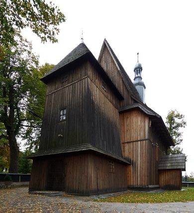 Zdjęcie nr 5: Kościół drewniany, orientowany, usytuowany na wzgórku, otoczony pierwotnym cmentarzem przykościelnym oraz starodrzewiem (lipami), ujęty drewnianym ogrodzeniem z poziomo ułożonych desek, w którego ciągu znajduje się masywna dzwonnica. Ściany kościoła wzniesione w konstrukcji zrębowej, oszalowanej pionowo z listwowaniem. Nawa na planie prostokąta; prezbiterium węższe, zamknięte wielobocznie, zbudowane na zrąb; od północy zakrystia na planie prostokąta; od południa późniejsza, dobudowana kruchta. Od zachodu nawę poprzedza niższa wieża, zbudowana w konstrukcji słupowo-ramowej, o pochyłych ścianach. Wnętrze nakryte stropami, w nawie z zaskrzynieniami. Nawa od prezbiterium oddzielona tęczą o łuku półkolistym wspartym na kroksztynach; na belce umieszczone rzeźby z początku XVI wieku przedstawiające grupę Ukrzyżowania: krucyfiks, Matka Boska, św. Jan Ewangelista oraz św. Maria Magdalena. Chór muzyczny wsparty na dwóch kolumnach, barokowy. Wnętrze pokryte polichromią z pierwszej połowy XVII wieku. Pośrodku kościoła oraz bliżej chóru muzycznego znajdują się wejścia do krypt pokryte płytami. Główny portal kościoła umieszczony jest od strony zachodniej, w przedsionku pod wieżą, ukształtowany w formie profilowanej, lekko zaostrzonej arkady przechodzącej w tzw. ośli grzbiet. Portal południowy, w kruchcie zakończony ostrołukiem, z motywem sznura w profilowaniach z pierwotnymi jednoskrzydłowymi drzwiami o gotyckich okuciach. Portal prowadzący z prezbiterium do zakrystii ma bogate profilowanie (zewnętrzny profil z motywem „ząbków”), jest zakończony w tzw. ośli grzbiet z drzwiami żelaznymi, dekorowanymi ukośną kratą. Okna znajdują się od strony południowej, pod dwa w nawie oraz w prezbiterium. Nadokienniki mają wykrój w ośli grzbiet. Ponadto jedno okno o analogicznym układzie w prezbiterium na ścianie wschodniej, częściowo zabudowane, zasłonięte ołtarzem. Dach gontowy o jednej kalenicy, wsparty na kroksztynach z szerokim okapem, o dużym kącie spadku uzyskanym poprzez zastosowaną gotycką więźbę storczykową, pośrodku z wieżyczką na sygnaturkę o barkowym kształcie. Wieża główna nakryta dachem gontowym, trójspadowym.
