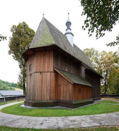 Zdjęcie nr 8: Kościół drewniany, orientowany, usytuowany na wzgórku, otoczony pierwotnym cmentarzem przykościelnym oraz starodrzewiem (lipami), ujęty drewnianym ogrodzeniem z poziomo ułożonych desek, w którego ciągu znajduje się masywna dzwonnica. Ściany kościoła wzniesione w konstrukcji zrębowej, oszalowanej pionowo z listwowaniem. Nawa na planie prostokąta; prezbiterium węższe, zamknięte wielobocznie, zbudowane na zrąb; od północy zakrystia na planie prostokąta; od południa późniejsza, dobudowana kruchta. Od zachodu nawę poprzedza niższa wieża, zbudowana w konstrukcji słupowo-ramowej, o pochyłych ścianach. Wnętrze nakryte stropami, w nawie z zaskrzynieniami. Nawa od prezbiterium oddzielona tęczą o łuku półkolistym wspartym na kroksztynach; na belce umieszczone rzeźby z początku XVI wieku przedstawiające grupę Ukrzyżowania: krucyfiks, Matka Boska, św. Jan Ewangelista oraz św. Maria Magdalena. Chór muzyczny wsparty na dwóch kolumnach, barokowy. Wnętrze pokryte polichromią z pierwszej połowy XVII wieku. Pośrodku kościoła oraz bliżej chóru muzycznego znajdują się wejścia do krypt pokryte płytami. Główny portal kościoła umieszczony jest od strony zachodniej, w przedsionku pod wieżą, ukształtowany w formie profilowanej, lekko zaostrzonej arkady przechodzącej w tzw. ośli grzbiet. Portal południowy, w kruchcie zakończony ostrołukiem, z motywem sznura w profilowaniach z pierwotnymi jednoskrzydłowymi drzwiami o gotyckich okuciach. Portal prowadzący z prezbiterium do zakrystii ma bogate profilowanie (zewnętrzny profil z motywem „ząbków”), jest zakończony w tzw. ośli grzbiet z drzwiami żelaznymi, dekorowanymi ukośną kratą. Okna znajdują się od strony południowej, pod dwa w nawie oraz w prezbiterium. Nadokienniki mają wykrój w ośli grzbiet. Ponadto jedno okno o analogicznym układzie w prezbiterium na ścianie wschodniej, częściowo zabudowane, zasłonięte ołtarzem. Dach gontowy o jednej kalenicy, wsparty na kroksztynach z szerokim okapem, o dużym kącie spadku uzyskanym poprzez zastosowaną gotycką więźbę storczykową, pośrodku z wieżyczką na sygnaturkę o barkowym kształcie. Wieża główna nakryta dachem gontowym, trójspadowym.
