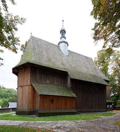 Zdjęcie nr 7: Kościół drewniany, orientowany, usytuowany na wzgórku, otoczony pierwotnym cmentarzem przykościelnym oraz starodrzewiem (lipami), ujęty drewnianym ogrodzeniem z poziomo ułożonych desek, w którego ciągu znajduje się masywna dzwonnica. Ściany kościoła wzniesione w konstrukcji zrębowej, oszalowanej pionowo z listwowaniem. Nawa na planie prostokąta; prezbiterium węższe, zamknięte wielobocznie, zbudowane na zrąb; od północy zakrystia na planie prostokąta; od południa późniejsza, dobudowana kruchta. Od zachodu nawę poprzedza niższa wieża, zbudowana w konstrukcji słupowo-ramowej, o pochyłych ścianach. Wnętrze nakryte stropami, w nawie z zaskrzynieniami. Nawa od prezbiterium oddzielona tęczą o łuku półkolistym wspartym na kroksztynach; na belce umieszczone rzeźby z początku XVI wieku przedstawiające grupę Ukrzyżowania: krucyfiks, Matka Boska, św. Jan Ewangelista oraz św. Maria Magdalena. Chór muzyczny wsparty na dwóch kolumnach, barokowy. Wnętrze pokryte polichromią z pierwszej połowy XVII wieku. Pośrodku kościoła oraz bliżej chóru muzycznego znajdują się wejścia do krypt pokryte płytami. Główny portal kościoła umieszczony jest od strony zachodniej, w przedsionku pod wieżą, ukształtowany w formie profilowanej, lekko zaostrzonej arkady przechodzącej w tzw. ośli grzbiet. Portal południowy, w kruchcie zakończony ostrołukiem, z motywem sznura w profilowaniach z pierwotnymi jednoskrzydłowymi drzwiami o gotyckich okuciach. Portal prowadzący z prezbiterium do zakrystii ma bogate profilowanie (zewnętrzny profil z motywem „ząbków”), jest zakończony w tzw. ośli grzbiet z drzwiami żelaznymi, dekorowanymi ukośną kratą. Okna znajdują się od strony południowej, pod dwa w nawie oraz w prezbiterium. Nadokienniki mają wykrój w ośli grzbiet. Ponadto jedno okno o analogicznym układzie w prezbiterium na ścianie wschodniej, częściowo zabudowane, zasłonięte ołtarzem. Dach gontowy o jednej kalenicy, wsparty na kroksztynach z szerokim okapem, o dużym kącie spadku uzyskanym poprzez zastosowaną gotycką więźbę storczykową, pośrodku z wieżyczką na sygnaturkę o barkowym kształcie. Wieża główna nakryta dachem gontowym, trójspadowym.
