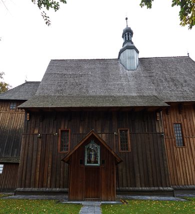 Zdjęcie nr 11: Kościół drewniany, orientowany, usytuowany na wzgórku, otoczony pierwotnym cmentarzem przykościelnym oraz starodrzewiem (lipami), ujęty drewnianym ogrodzeniem z poziomo ułożonych desek, w którego ciągu znajduje się masywna dzwonnica. Ściany kościoła wzniesione w konstrukcji zrębowej, oszalowanej pionowo z listwowaniem. Nawa na planie prostokąta; prezbiterium węższe, zamknięte wielobocznie, zbudowane na zrąb; od północy zakrystia na planie prostokąta; od południa późniejsza, dobudowana kruchta. Od zachodu nawę poprzedza niższa wieża, zbudowana w konstrukcji słupowo-ramowej, o pochyłych ścianach. Wnętrze nakryte stropami, w nawie z zaskrzynieniami. Nawa od prezbiterium oddzielona tęczą o łuku półkolistym wspartym na kroksztynach; na belce umieszczone rzeźby z początku XVI wieku przedstawiające grupę Ukrzyżowania: krucyfiks, Matka Boska, św. Jan Ewangelista oraz św. Maria Magdalena. Chór muzyczny wsparty na dwóch kolumnach, barokowy. Wnętrze pokryte polichromią z pierwszej połowy XVII wieku. Pośrodku kościoła oraz bliżej chóru muzycznego znajdują się wejścia do krypt pokryte płytami. Główny portal kościoła umieszczony jest od strony zachodniej, w przedsionku pod wieżą, ukształtowany w formie profilowanej, lekko zaostrzonej arkady przechodzącej w tzw. ośli grzbiet. Portal południowy, w kruchcie zakończony ostrołukiem, z motywem sznura w profilowaniach z pierwotnymi jednoskrzydłowymi drzwiami o gotyckich okuciach. Portal prowadzący z prezbiterium do zakrystii ma bogate profilowanie (zewnętrzny profil z motywem „ząbków”), jest zakończony w tzw. ośli grzbiet z drzwiami żelaznymi, dekorowanymi ukośną kratą. Okna znajdują się od strony południowej, pod dwa w nawie oraz w prezbiterium. Nadokienniki mają wykrój w ośli grzbiet. Ponadto jedno okno o analogicznym układzie w prezbiterium na ścianie wschodniej, częściowo zabudowane, zasłonięte ołtarzem. Dach gontowy o jednej kalenicy, wsparty na kroksztynach z szerokim okapem, o dużym kącie spadku uzyskanym poprzez zastosowaną gotycką więźbę storczykową, pośrodku z wieżyczką na sygnaturkę o barkowym kształcie. Wieża główna nakryta dachem gontowym, trójspadowym.
